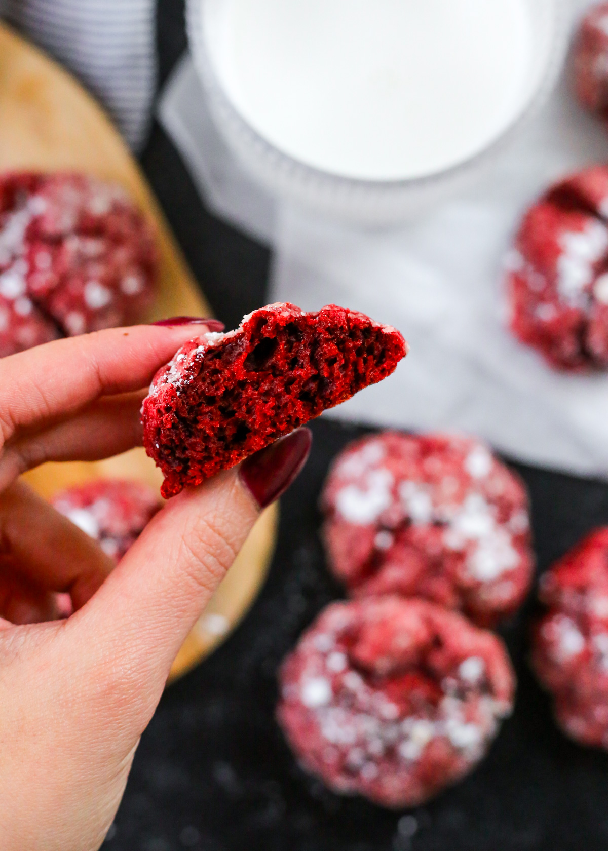A woman's hand holds up a red velvet gooey butter cookie that's been folded and torn in half to display the inside of the cookie, which is vibrant red with a moist, cake-like consistency. The remaining cookies are visible in the background along with a glass of milk