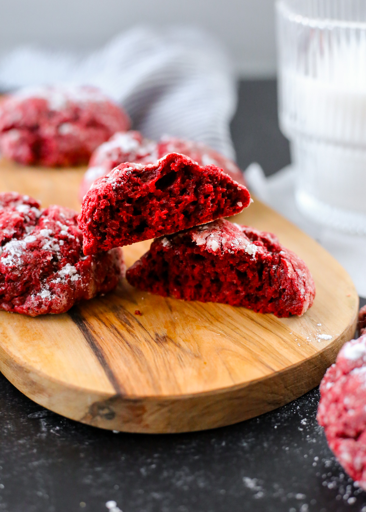 A red velvet gooey butter cookie is displayed on a small wooden serving board, broken open and facing the two halves towards the camera to show the interior of the cookie which is bright red and crumbly with a moist, cake-like texture
