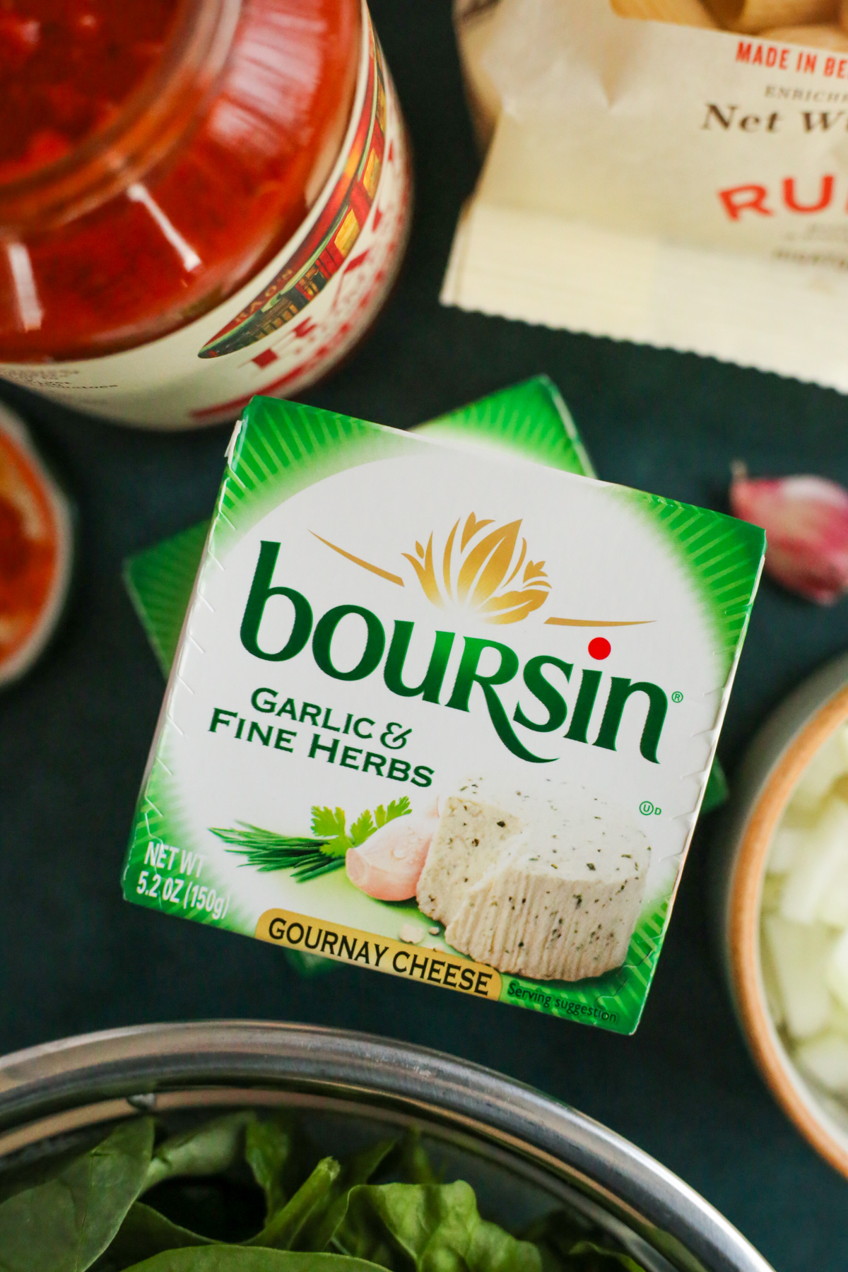 A closer view of the packaging for Boursin cheese in the Garlic and Fine Herbs flavor, stacked on top of another package and displayed alongside other ingredients on a dark teal green backdrop