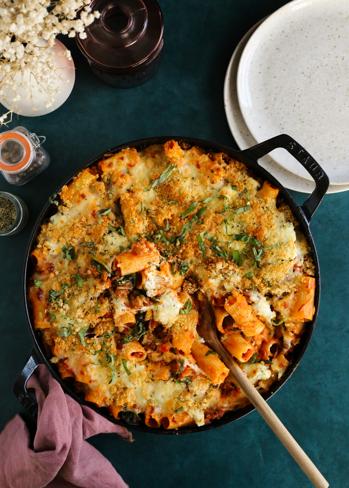 An overhead view of a round casserole dish on a dark teal backdrop, filled with a baked rigatoni recipe featuring a tomato sauce, fresh basil, and melted cheese, with a wooden serving spoon scooping out of the middle to reveal the melty cheese