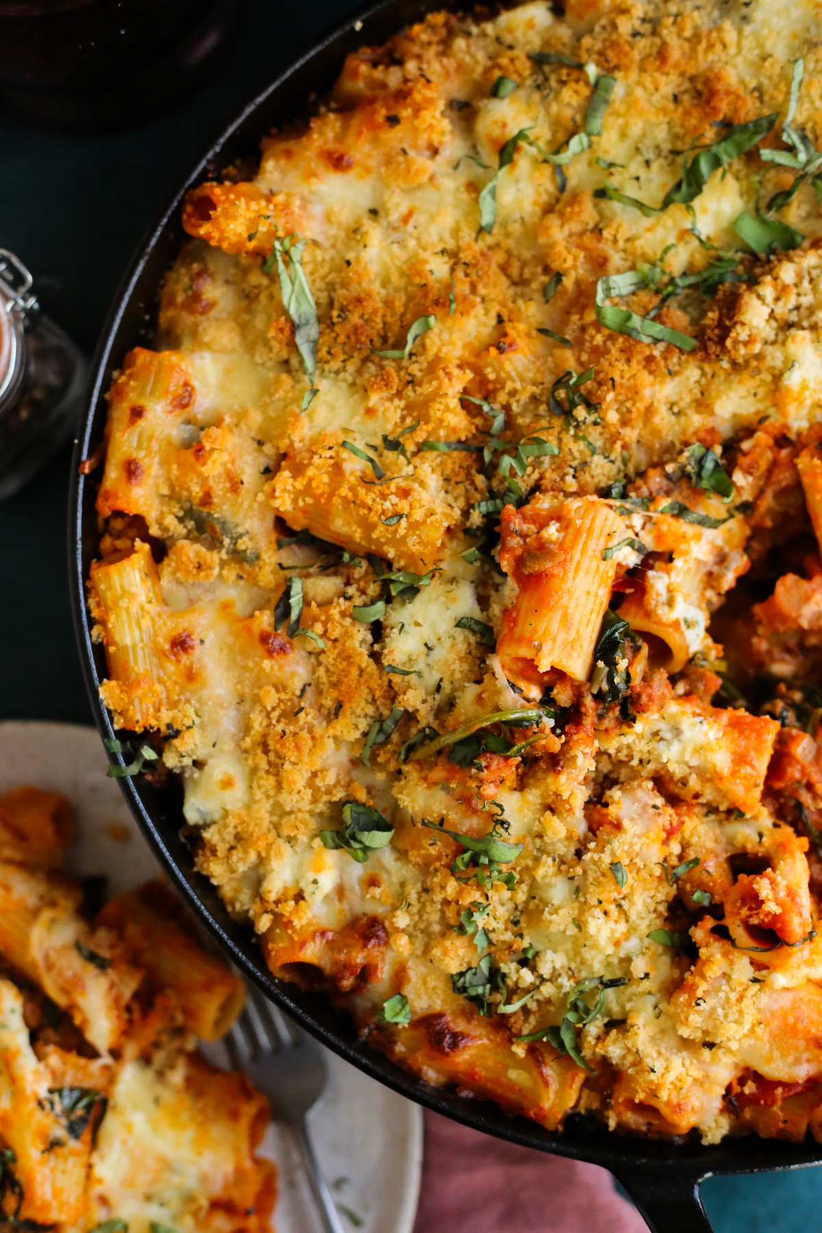 Overhead view into a round casserole dish filled with a baked rigatoni recipe, with a melted cheese and breadcrumb topping and some fresh basil  on top