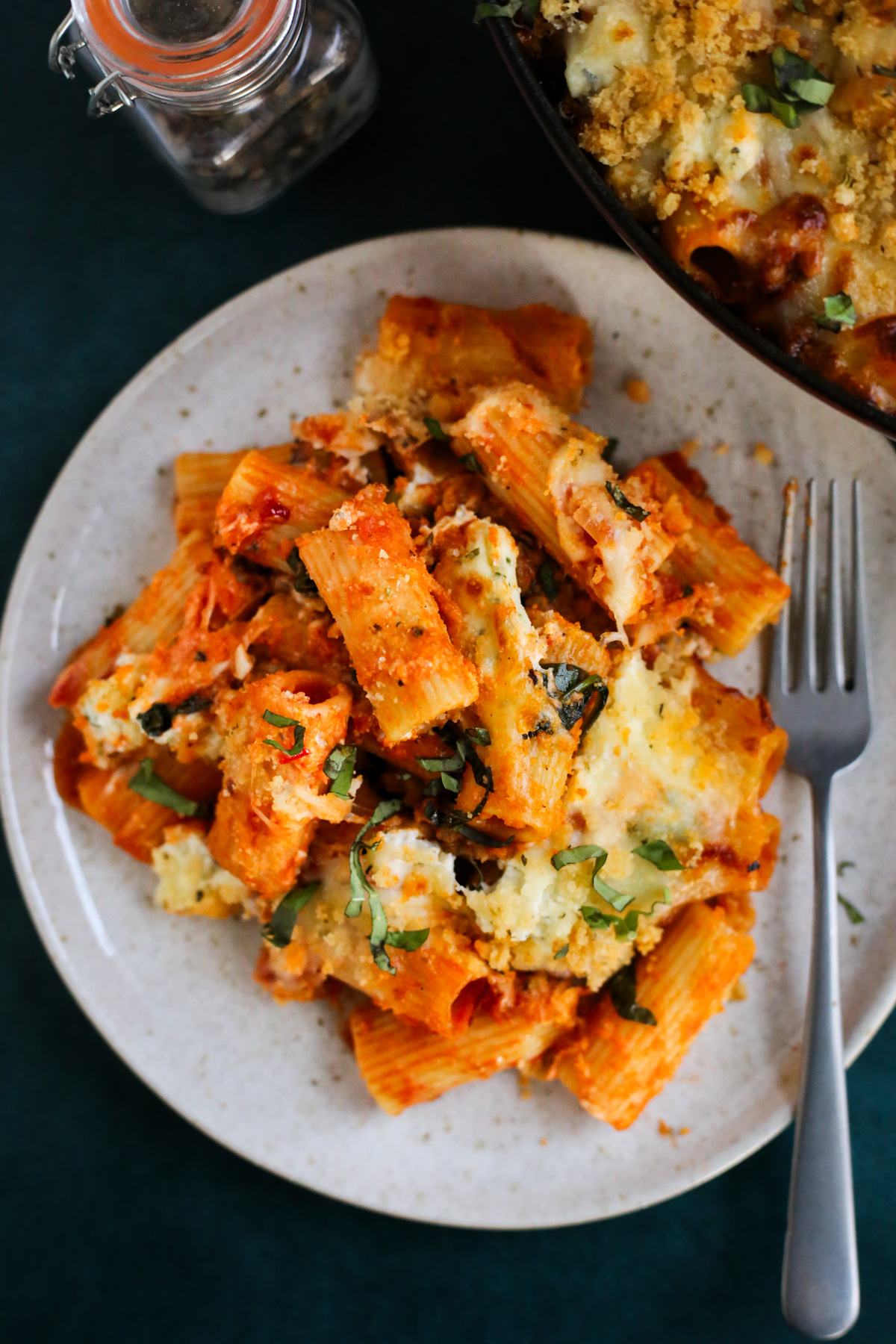 Overhead view of a dinner plate serving a baked rigatoni recipe that's garnished with melty cheese, breadcrumbs, and fresh basil, with everything coated in a rich tomato sauce and a silver spoon resting on the right side of the dinner plate