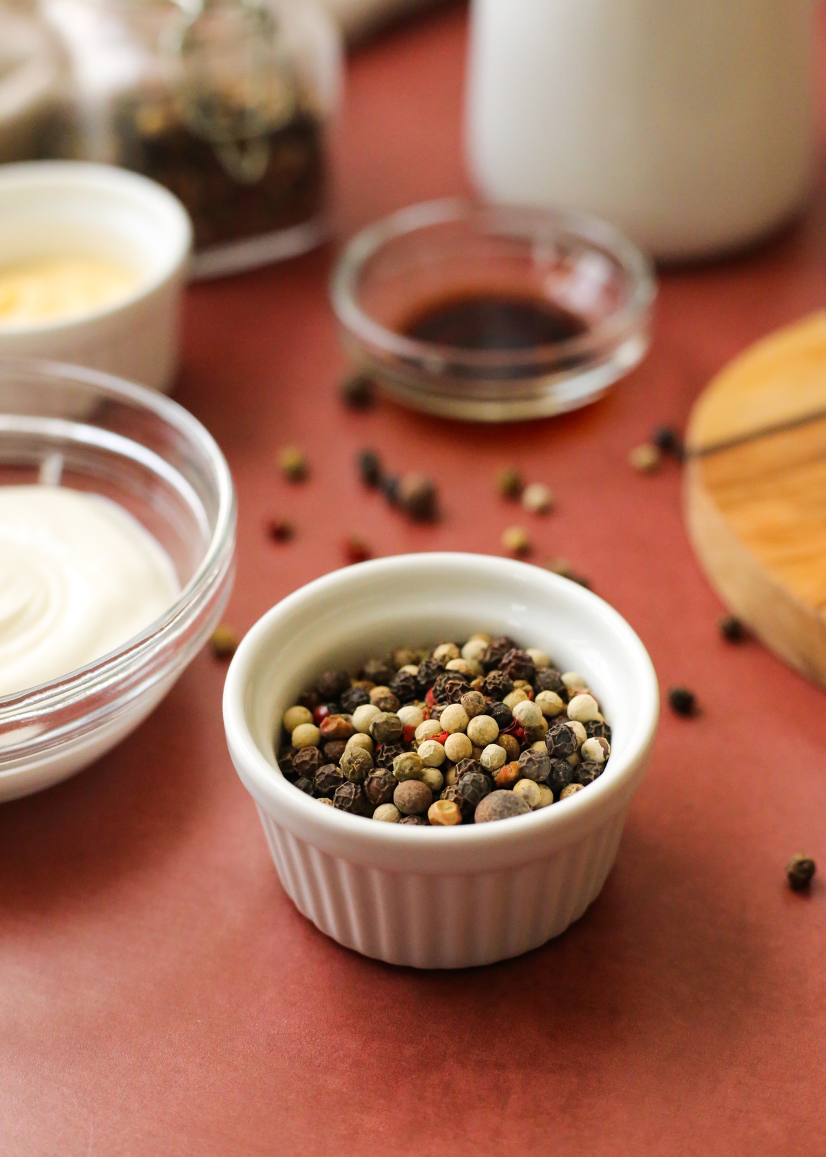 An angled view into a small white ramekin filled with whole peppercorns in varying shades of brown, white, and red, with additional ingredients to make a creamy peppercorn dressing in other small prep bowls in the background