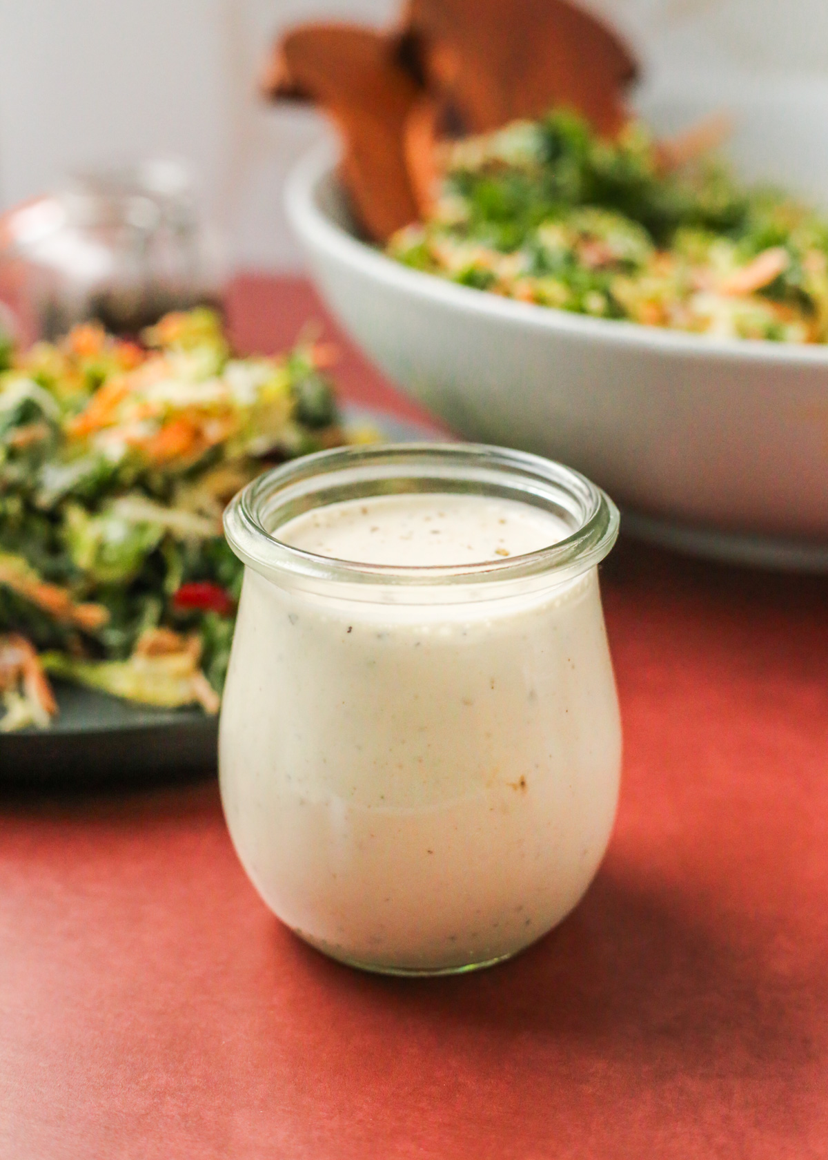 A clear, teardrop shaped glass jar filled to the brim with a homemade creamy peppercorn salad dressing, with fleck of cracked pepper visible throughout, served in front of a plated side salad coated with the dressing