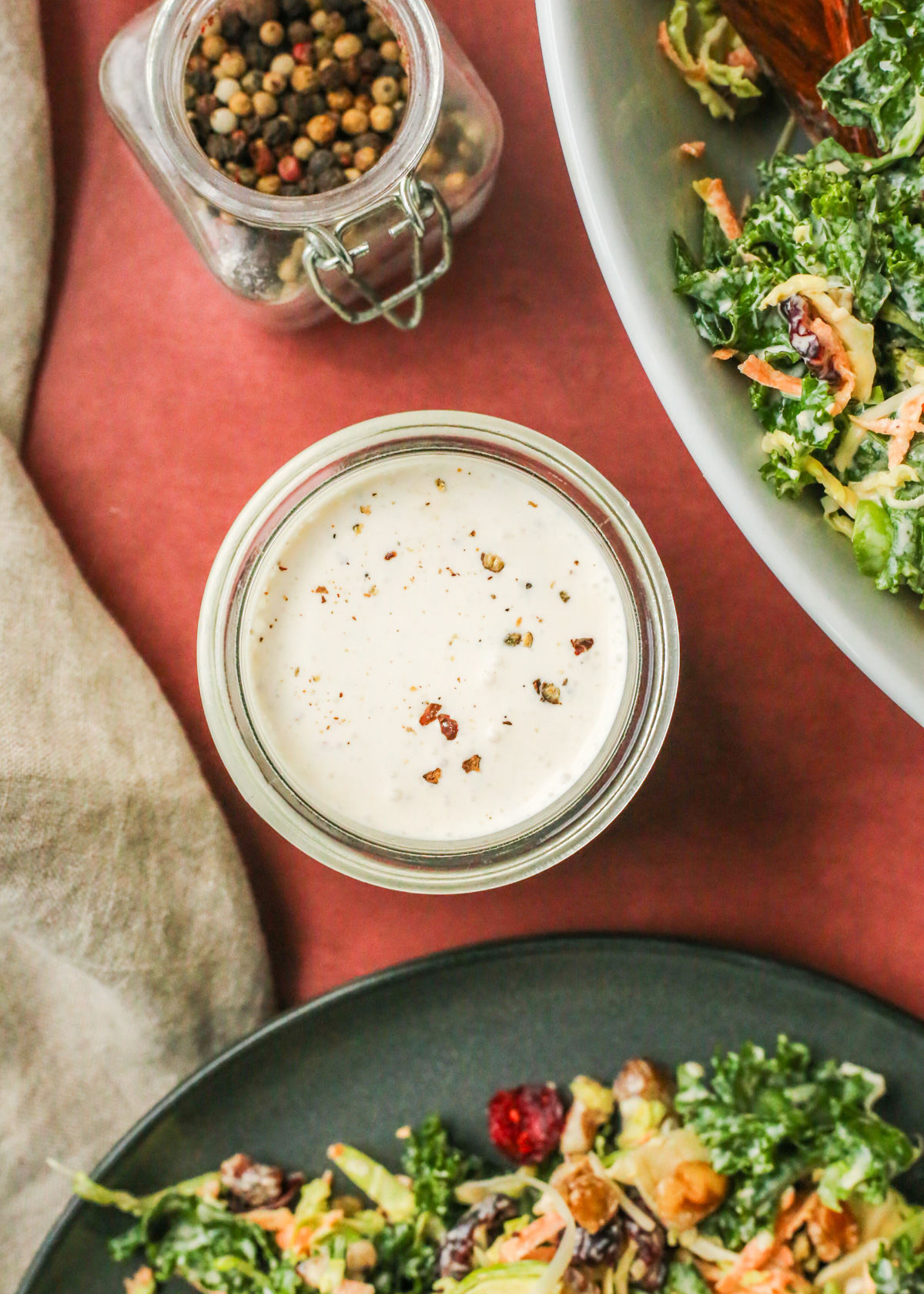 Overhead view of a small clear glass jar filled to the brim with a creamy peppercorn dressing, displayed alongside a small glass jar of whole peppercorns in various colors, and a green salad coated in the same dressing in the corners of the frame