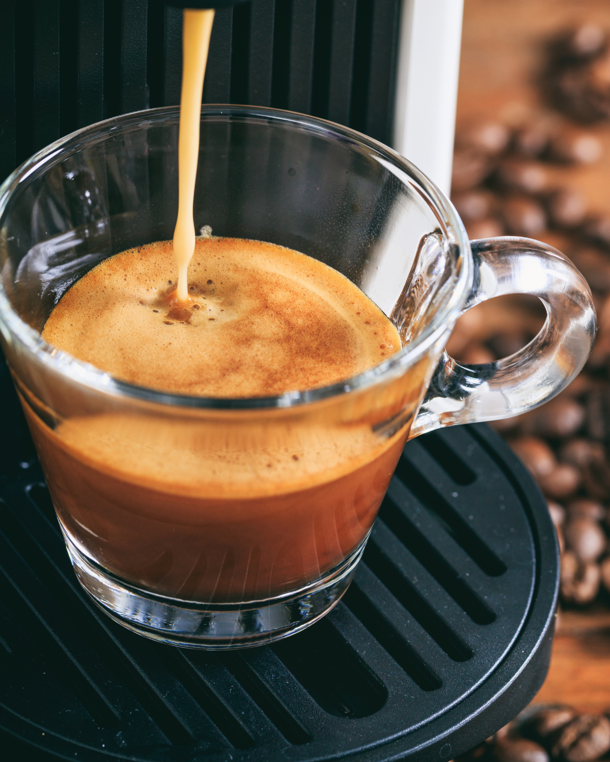 A view into a small glass handled espresso mug that is halfway filled with freshly brewed espresso, which is being dispensed from a black and silver espresso machine with whole coffee beans scattered in the background