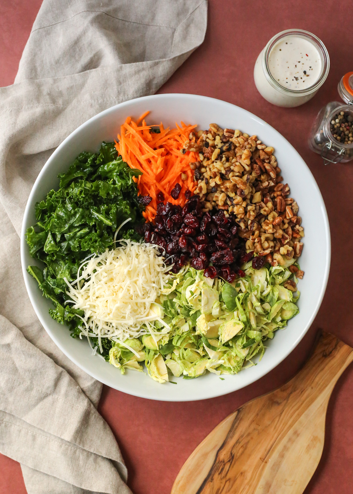 Overhead view into a large white mixing bowl on a kitchen countertop, filled with the unmixed ingredients for a kale and brussels sprout salad recipe, including massaged kale, shredded parmesan cheese, thinly sliced brussels sprouts, dried cranberries, chopped nuts, and shredded carrot, with a jar of creamy peppercorn dressing placed nearby