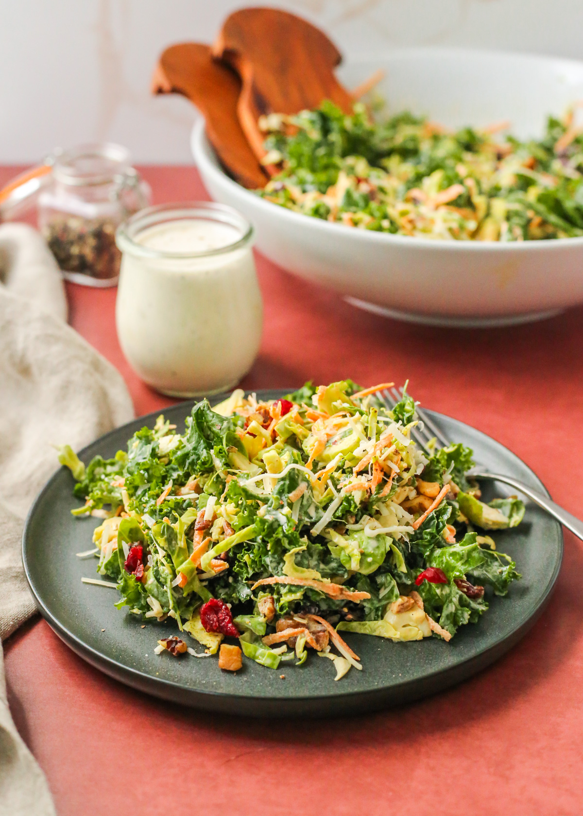A salad made from sliced brussels sprouts, massaged kale, shredded carrots, and dried cranberries plus walnuts and pecans is tossed with a creamy dressing and served on a dark ceramic plate in front of the remaining salad bowl and extra dressing