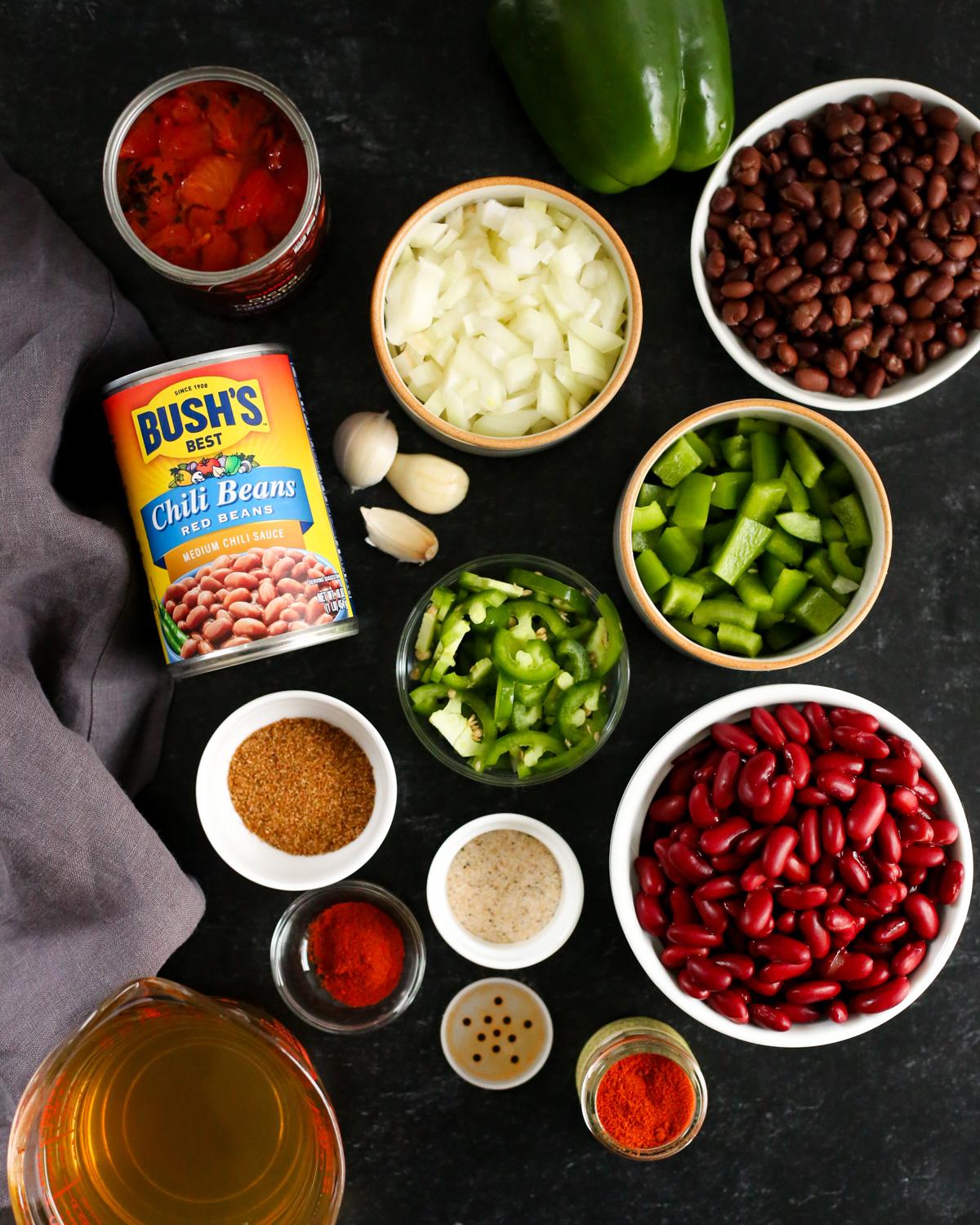 Overhead view of a collection of ingredients to make a chili recipe, including a can of Bush's Best chili beans, drained black beans and kidney beans, chopped onions, green bell peppers, jalapeño peppers, a few garlic cloves, a can of fire roasted tomatoes, and various chili spices and seasonings