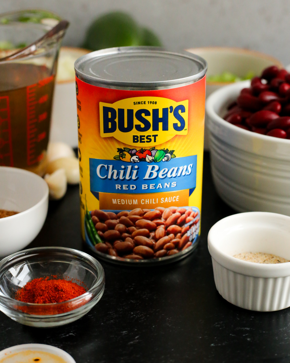 A can of Bush's Best Chili  Beans (red beans in a medium chili sauce) displayed on a dark countertop with various other chili seasonings and ramekins or prep bowls in the background