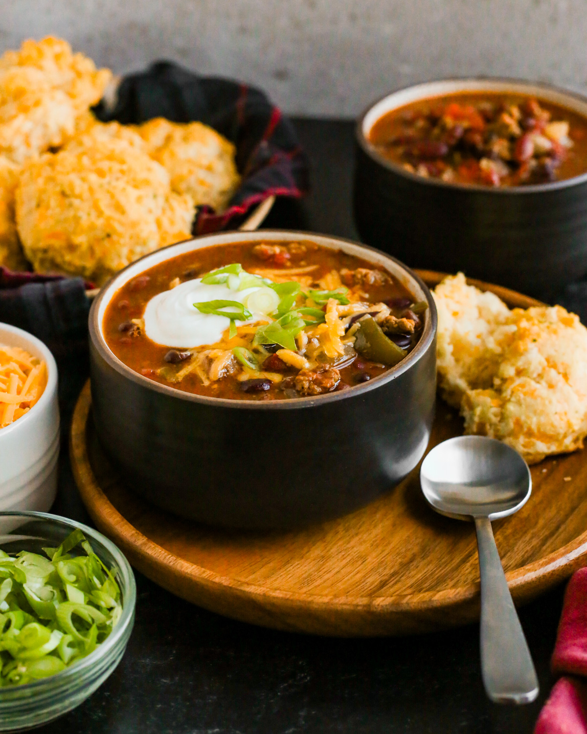 Styled image of a bowl of lamb chili, served in a kitchen scene with cheddar bay biscuits, shredded cheese, green onions, and a second bowl of chili in the background. The bowl in focus is topped with melted cheese, sour cream, and green onions and a soup spoon rests on the wooden plate it's served on