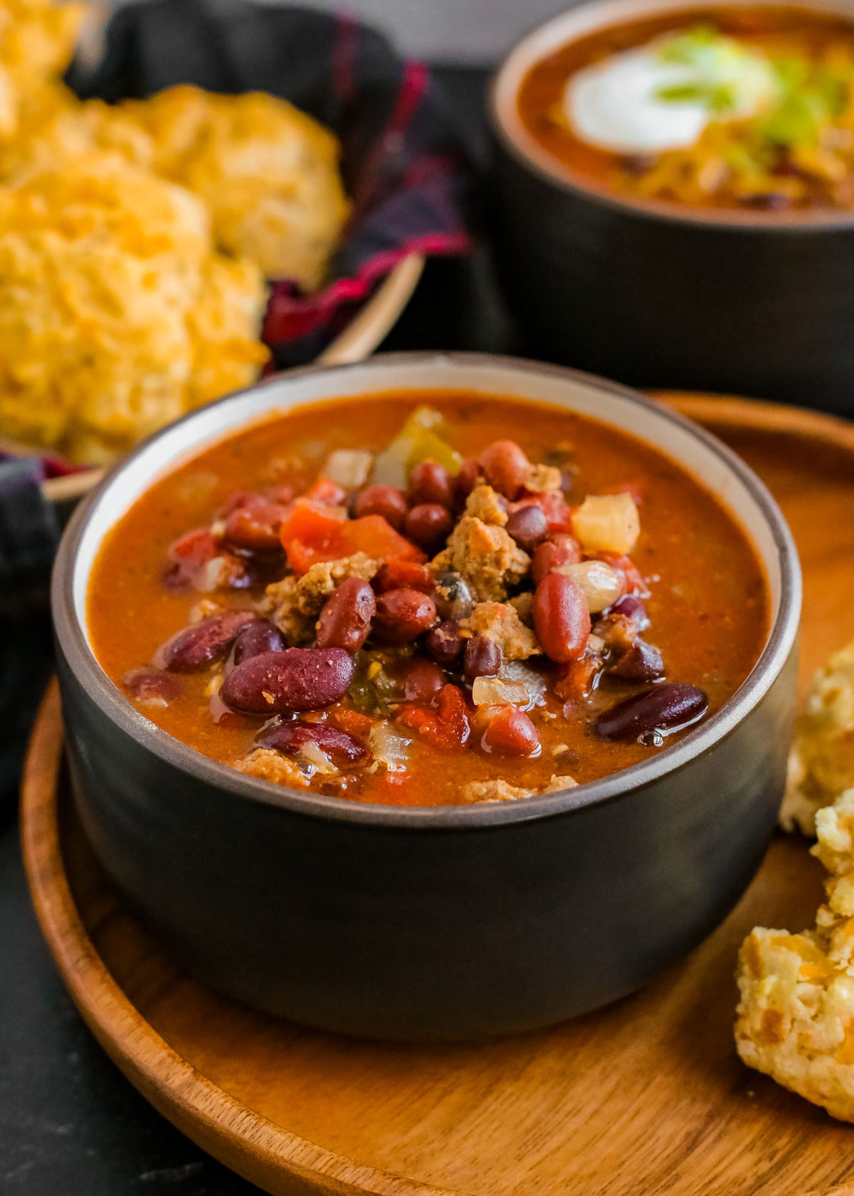 A dark ceramic serving bowl filled to the brim with a three bean lamb chili served on a wooden bowl with cheddar biscuits surrounding it. Pieces of tomato, onion, and ground lamb are visible in the rich, dark red stew