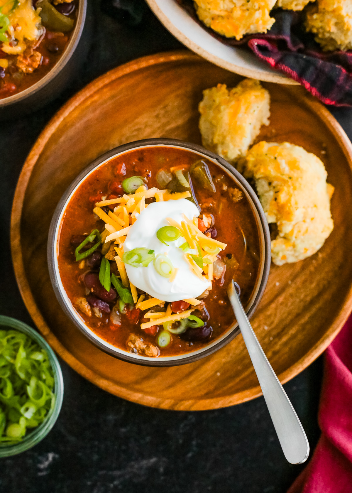 Overhead view of a bowl of lamb chili served on a wooden serving plate, topped with shredded cheddar cheese, a dollop of sour cream, and some sliced green onions with a soup spoon resting in the right side of the bowl