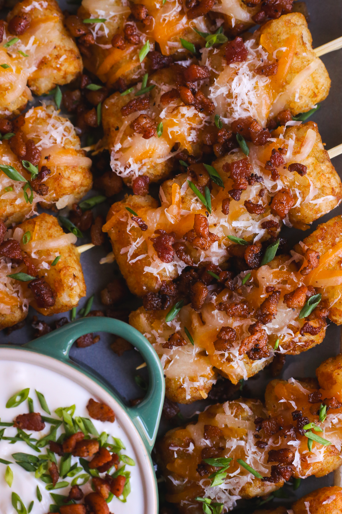 Close up view of loaded tater tots on skewers, with four tater tots stacked on each small wooden skewer, with melted cheese, crispy bacon bits, and chopped chives on top with a small ramekin holding a sour cream dip displayed on the same serving platter