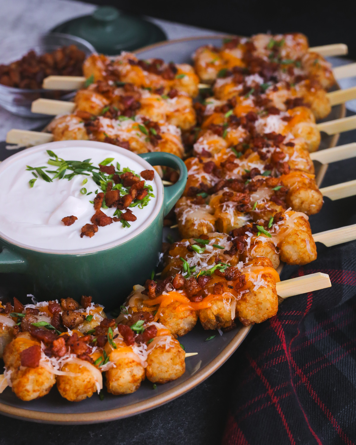A serving platter with rows of loaded tater tots on skewers, served with a sour cream dipping sauce topped with sliced green onions and crispy bacon bits