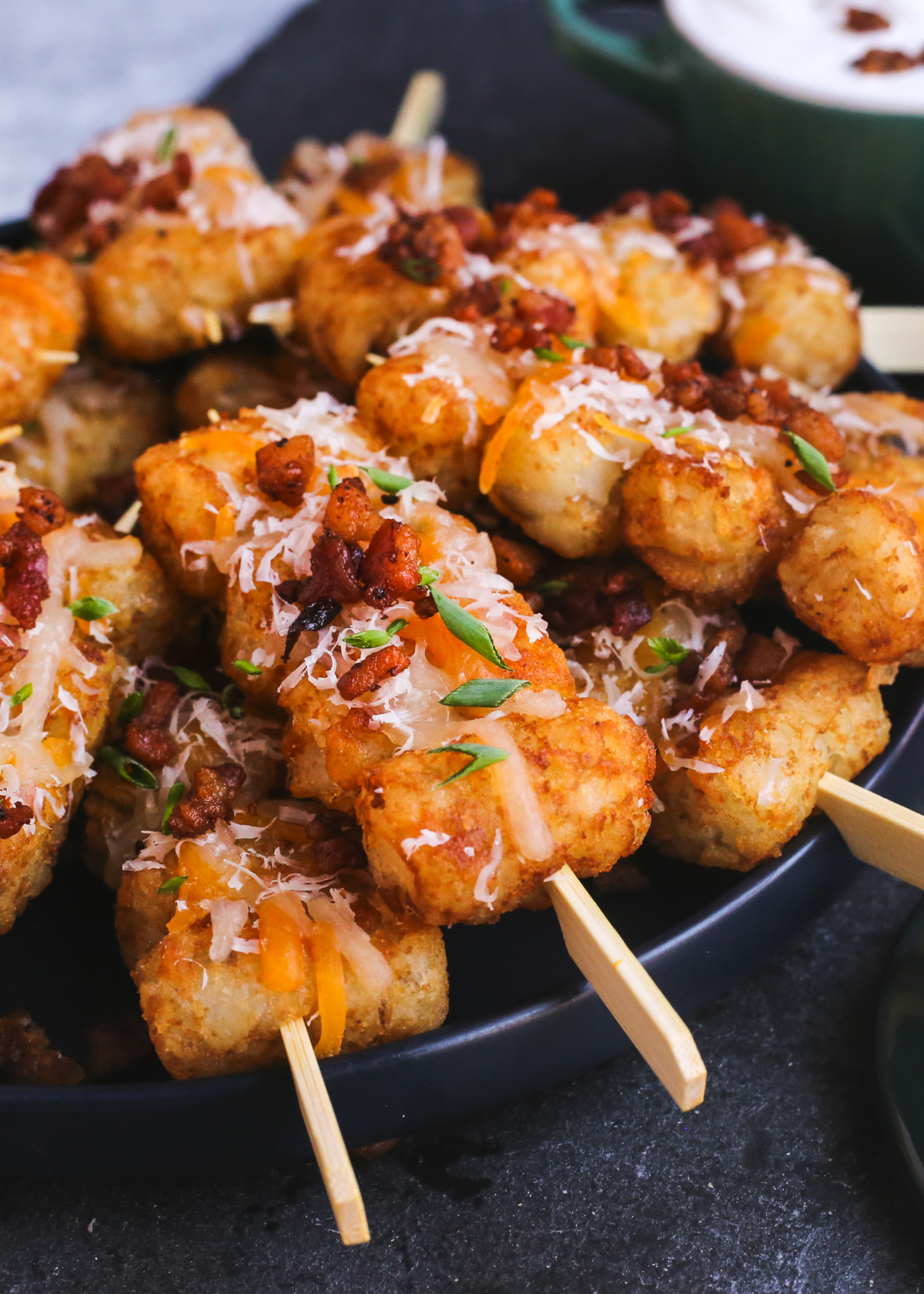 A stack of loaded tater tots on short wooden skewers is piled in a circular formation on a serving plate, with melted cheese, crispy bacon bits, and sliced green onions sprinkled over the top