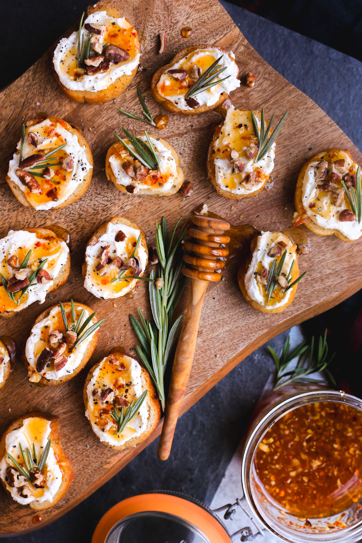 Overhead view of a wooden serving board with multiple whipped feta crostinis arranged on it, each topped with pecans, rosemary, and a drizzle of hot honey. A jar with leftover hot honey and extra sprigs of rosemary are placed nearby and a honey dipper coated with the hot honey is pictured on the serving board
