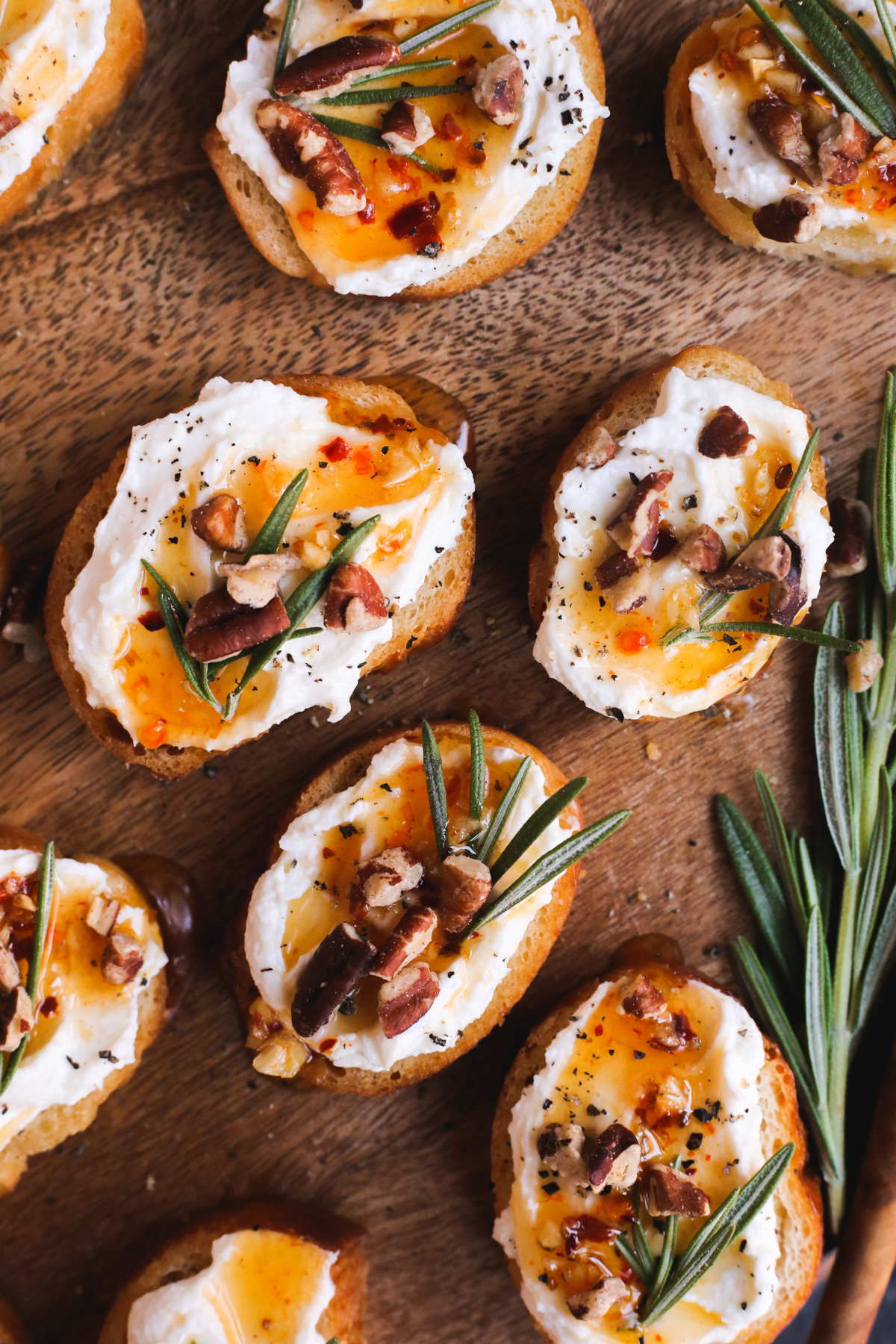 A small, round, toasted crostini is in the center of the photo, displayed on a wooden serving board with more crostinis surrounding it. It's garnished with chopped pecan pieces and fresh rosemary, which stand out against the white feta cheese. A hot honey drizzle and cracked black pepper is also included as a garnish