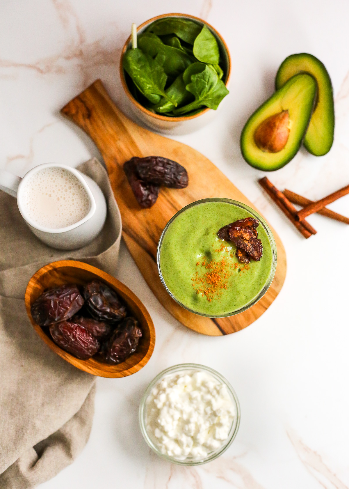 Overhead shot showing the ingredients needed to make a high protein green smoothie, including fresh dates, cottage cheese, milk, cinnamon sticks, avocado, and fresh spinach, with the blended smoothie centered in the middle of the frame, garnished with a few chopped dates and a dusting of cinnamon
