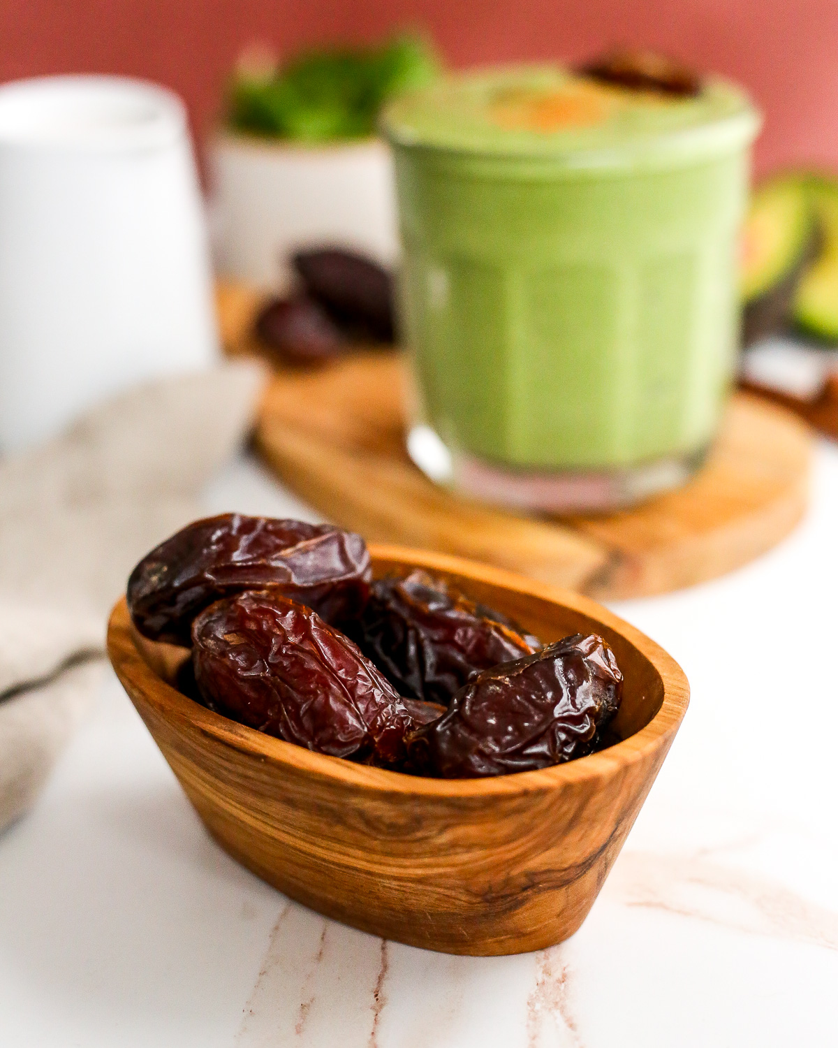 A focused shot of pitted Medjool dates, displayed in a small wooden prep bowl on a marble kitchen countertop with a green smoothie in the background