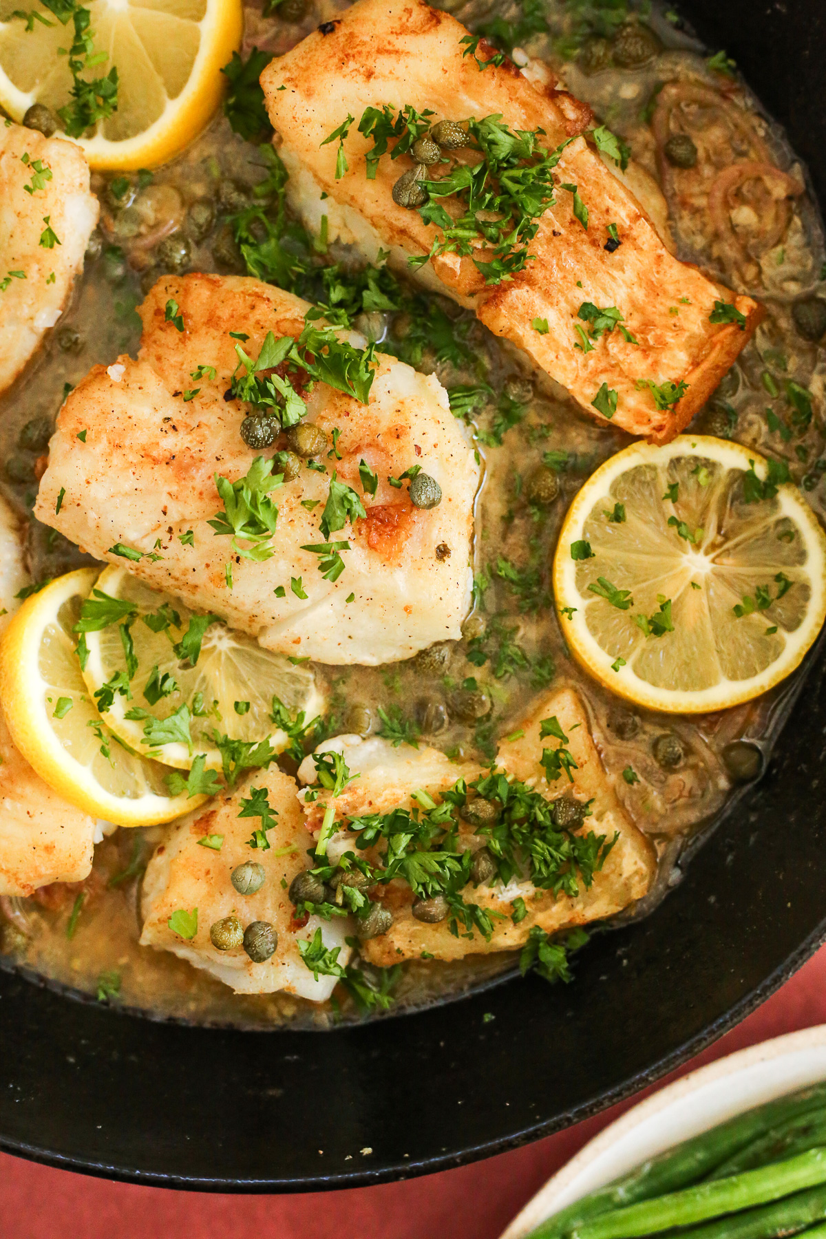 Overhead view of a cod piccata recipe in a dark cast iron braising pan, with lemon slices, capers, and chopped parsley arranged around and on top of a golden brown pan-seared cod fillet