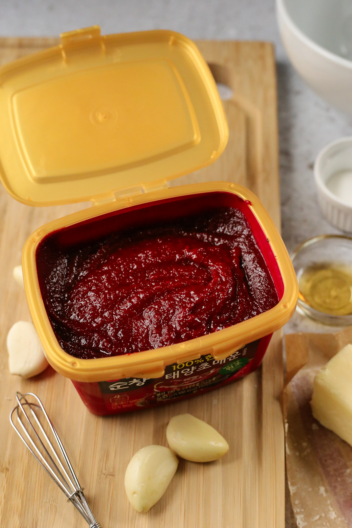 a view into the tub of gochujang used to make this recipe, which is nearly full of a thick, sticky, Korean red pepper paste, displayed on a wooden cutting board with a small whisk and several cloves of garlic.