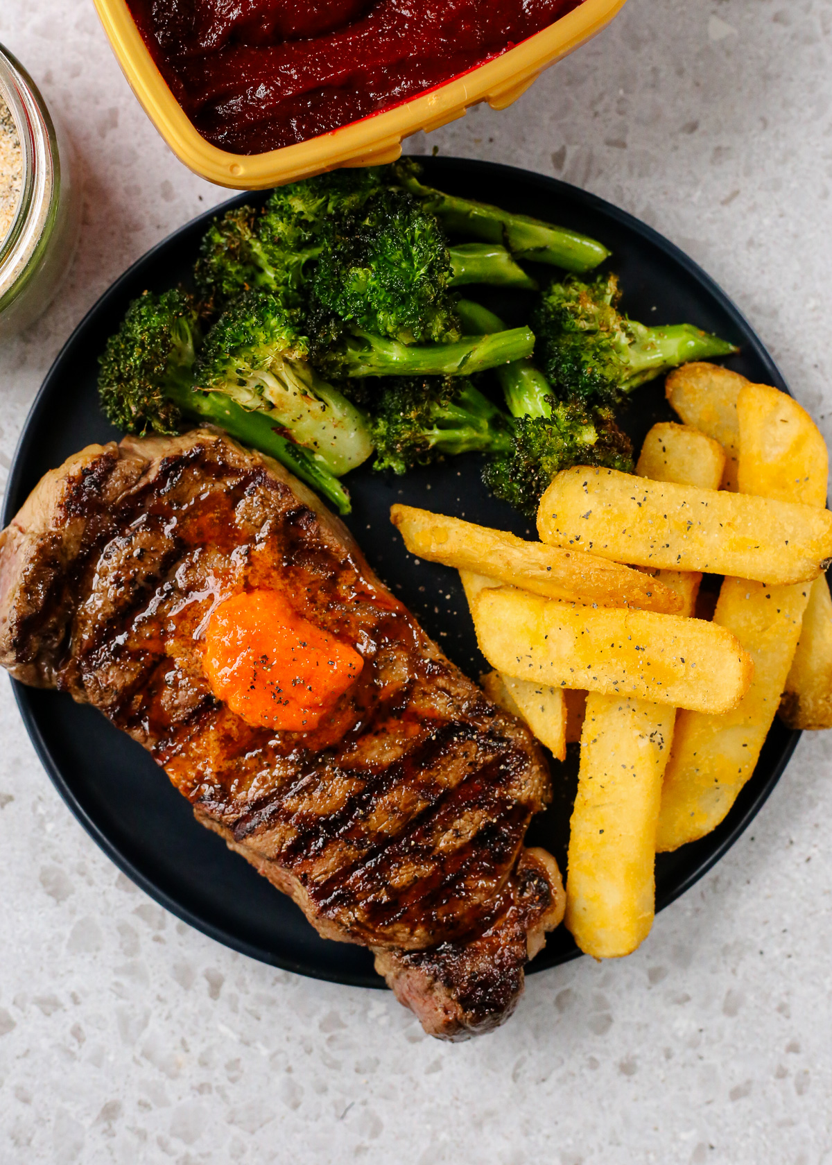 A large steak with prominent sear marks is served on a dark ceramic dinner plate with roasted broccoli and steak fries, topped with specks of black pepper and a dollop of bright orange gochujang butter, which is melting from the heat of the grilled steak.