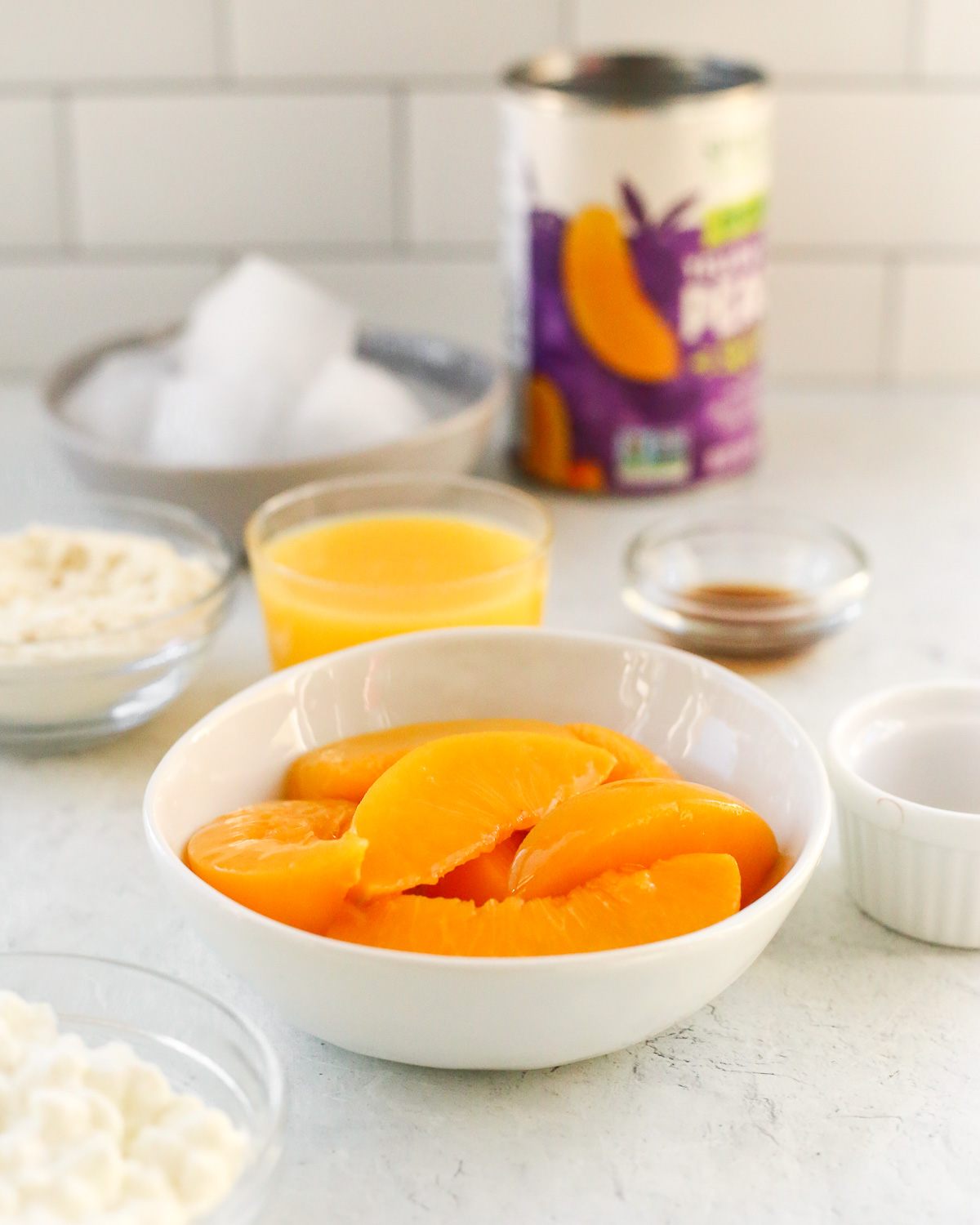 The ingredients to make a high protein peach smoothie recipe on a kitchen countertop, in small white or clear glass ramekins and prep bowls, including canned peaches, protein powder, cottage cheese, orange juice, vanilla extract, and ice cubes