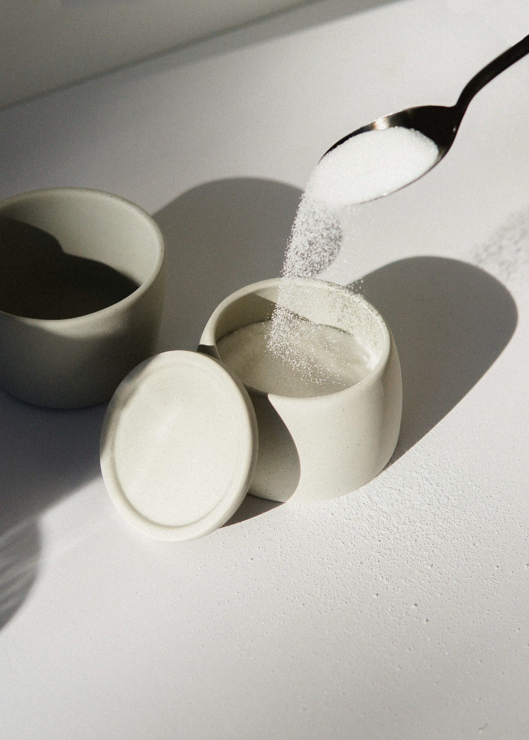 A spoon pouring a spoonful of sugar into a white ceramic dish with a lid propped against the side.
