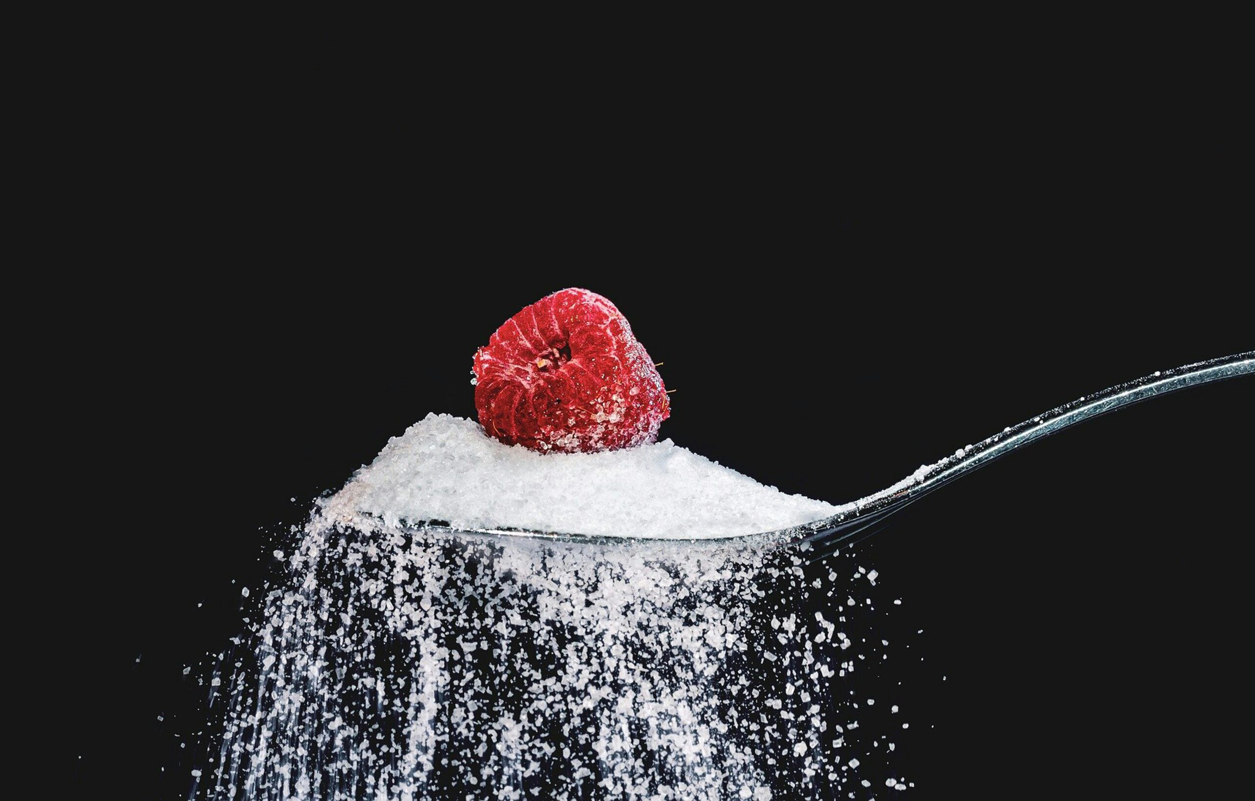 An overflowing spoonful of sugar with a fresh raspberry on top against a black background.