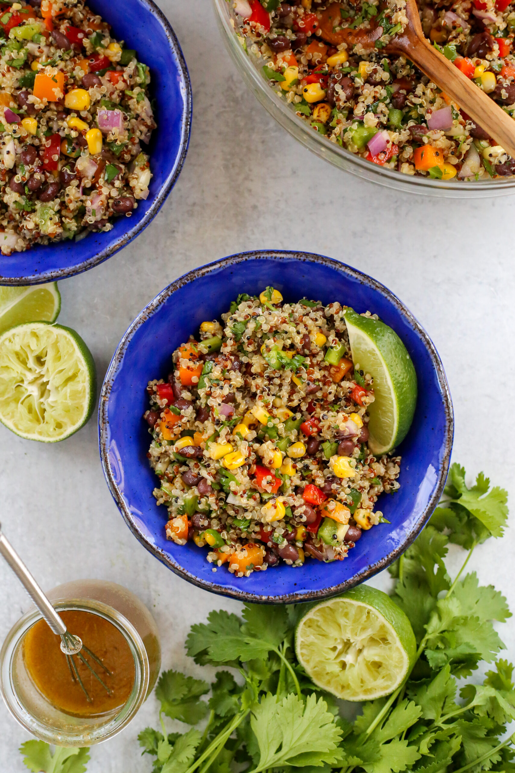 Overhead view of a bright blue bowl filled with a quinoa black bean salad, garnished with a lime wedge and surrounded by the mixing bowl it was served from and extra cumin lime dressing. 