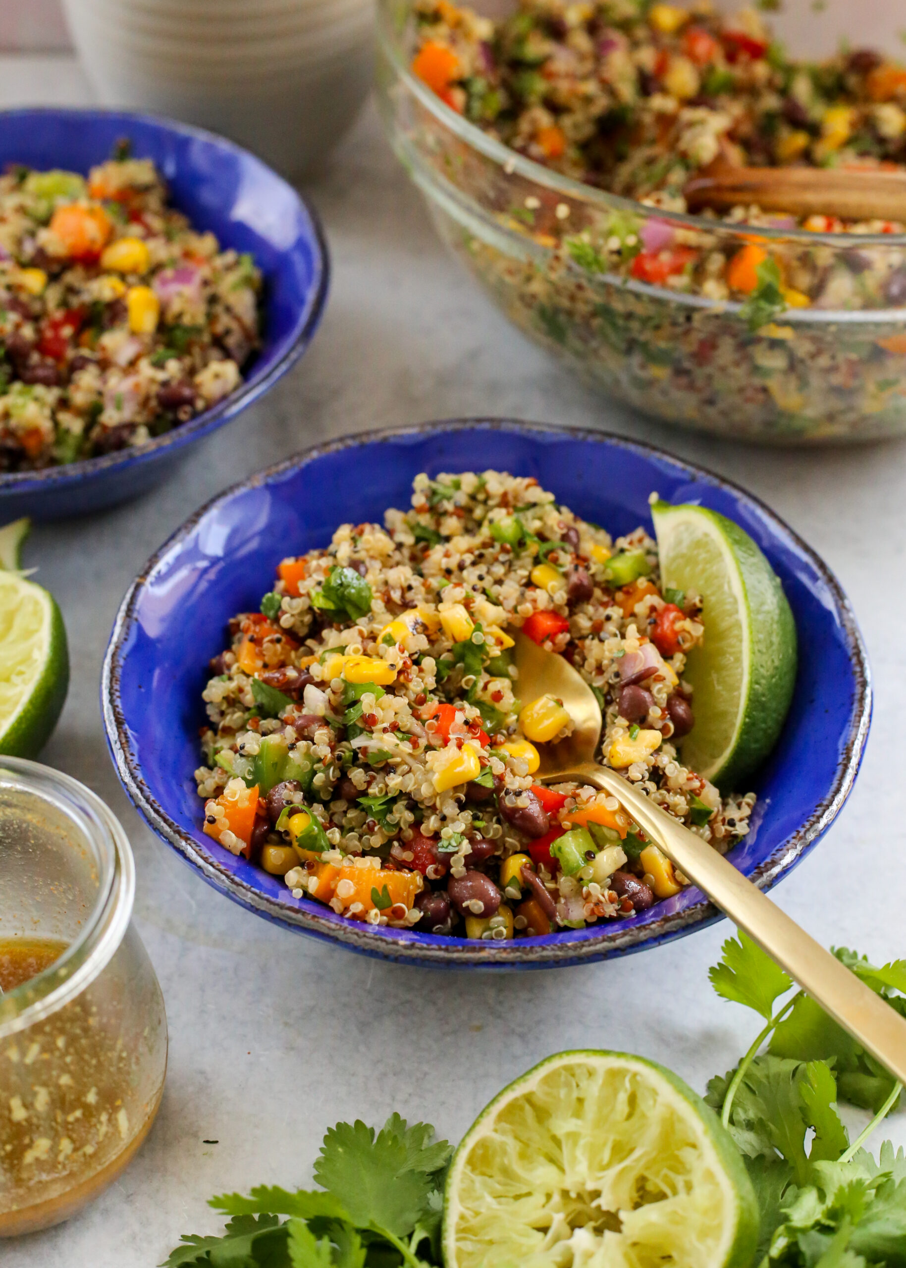 A bright blue ceramic bowl is full of a quinoa black bean salad featuring red, orange, and green bell pepper, black beans, red onions, sweet corn, and cilantro, garnished with a large lime wedge.