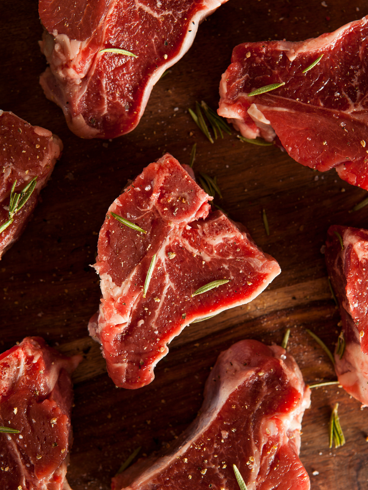 An arrangement of seasoned, uncooked lamb loin chops displayed on a cutting board, looking fresh and flavorful with salt, pepper, and bits of rosemary on top of each. 