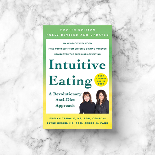 A copy of the 4th edition of the Intuitive Eating book rests on a white and grey marble countertop. 