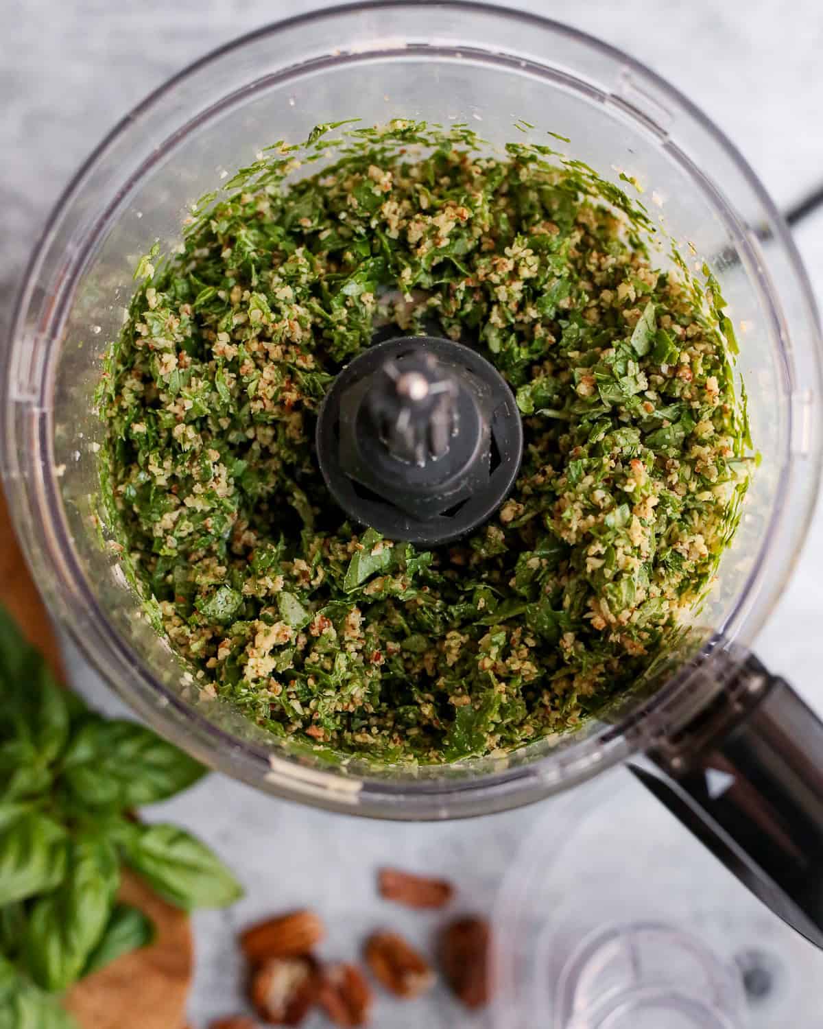 View into a food processor containing a roughly chopped mixture of basil and pecans. 