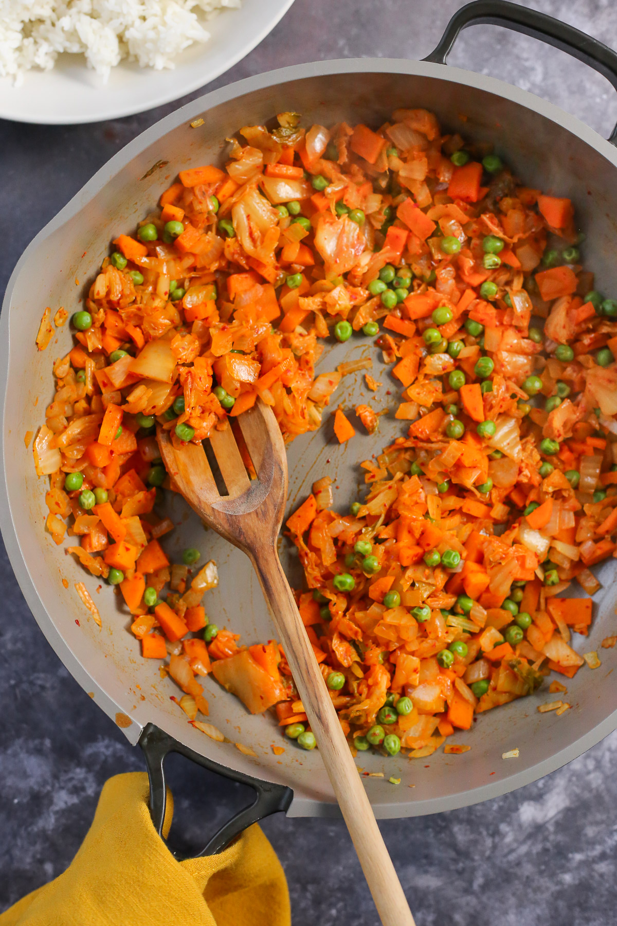 A stir-fried mixture of kimchi, carrots, onions, and green peas in a large skillet with a wooden spoon resting on the side. 