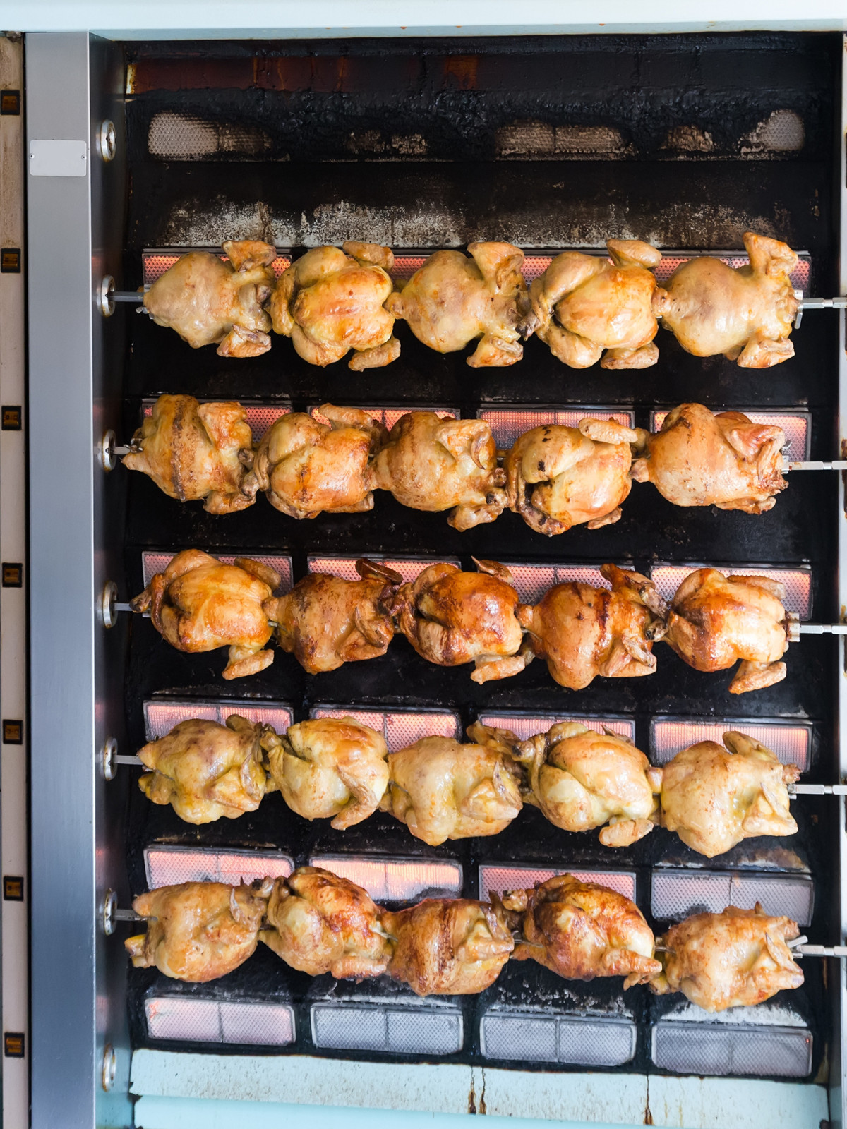 A commercial oven full of rotisserie chickens, rotating in front of a heat plate and seasoned with herbs. 