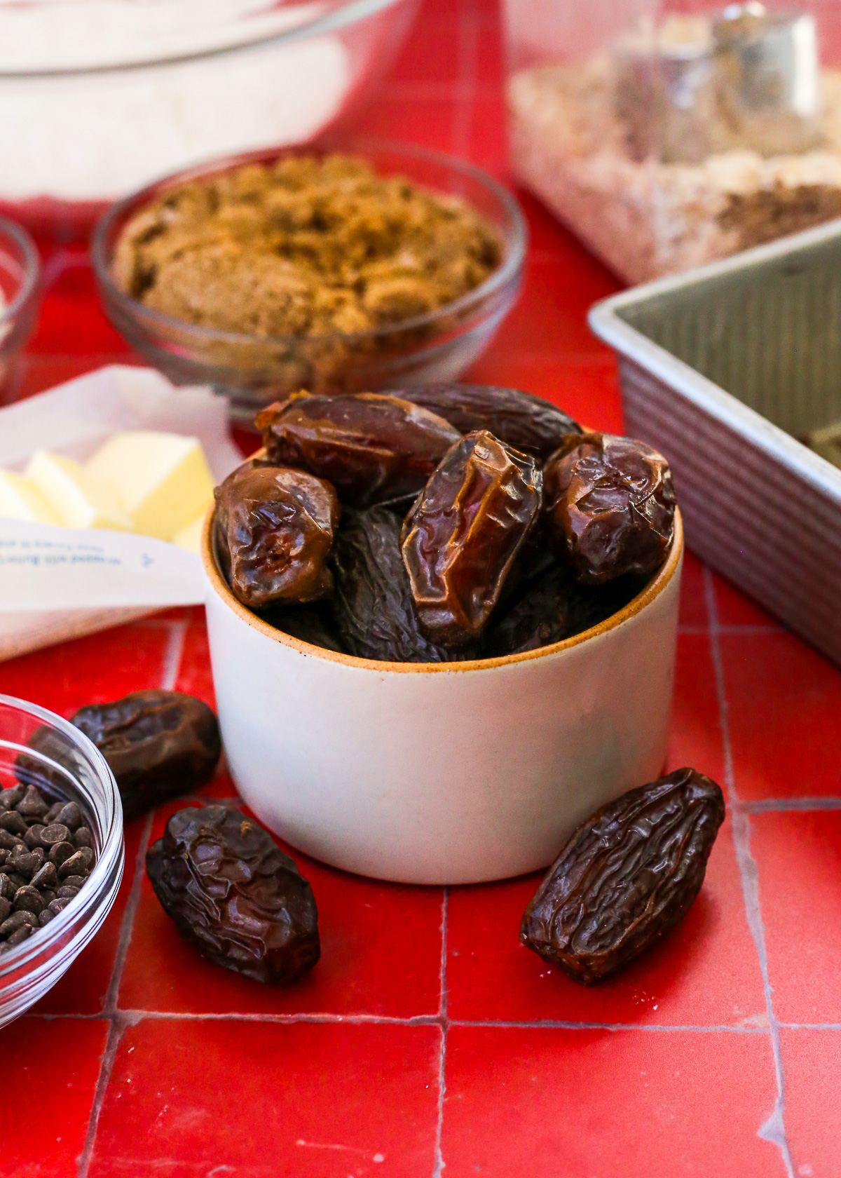 A small ceramic ramekin filled with dates, displayed on a red tile countertop with additional baking ingredients in the background.