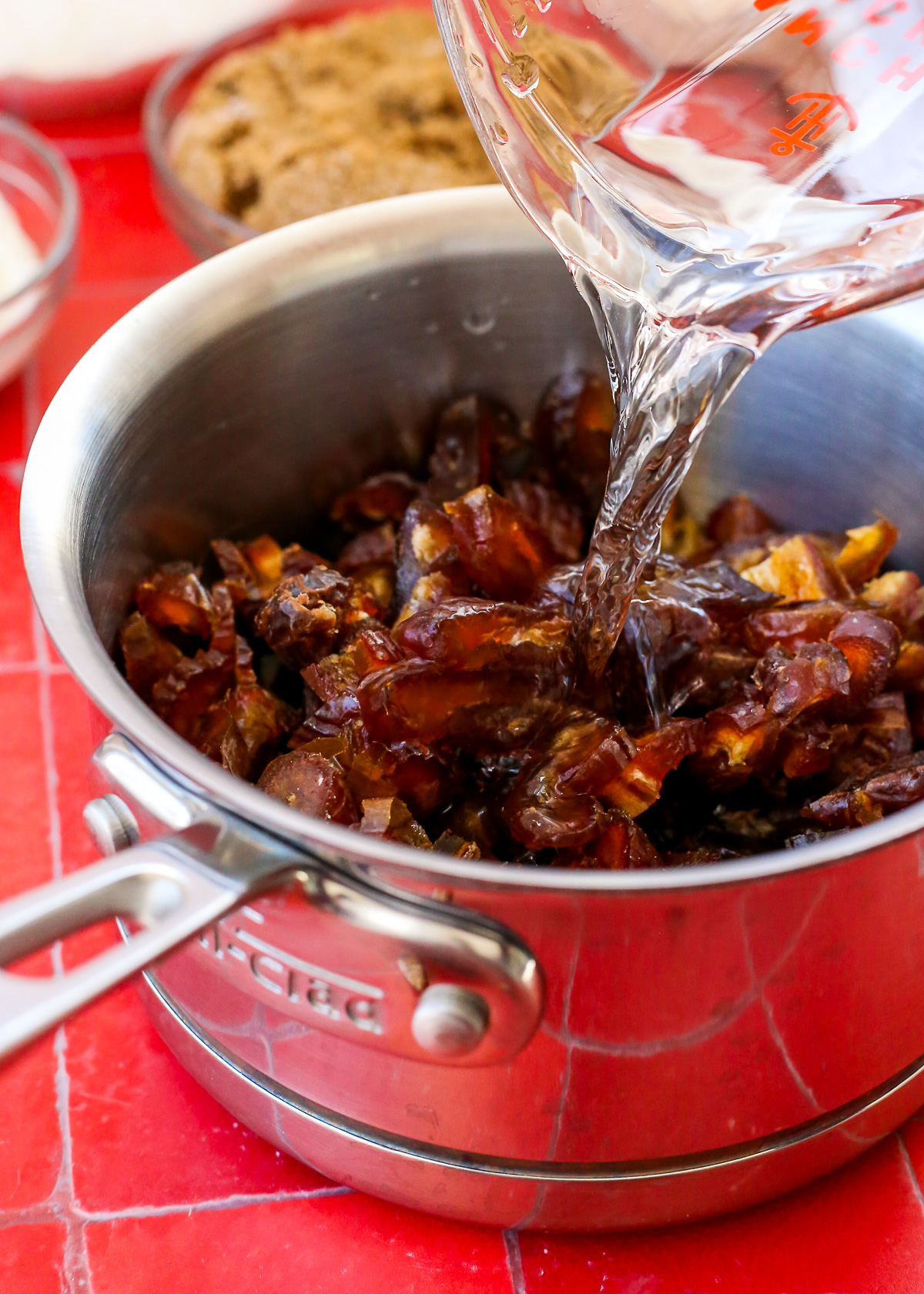 A clear measuring cup pours water into a saucepan filled with chopped pitted dates.