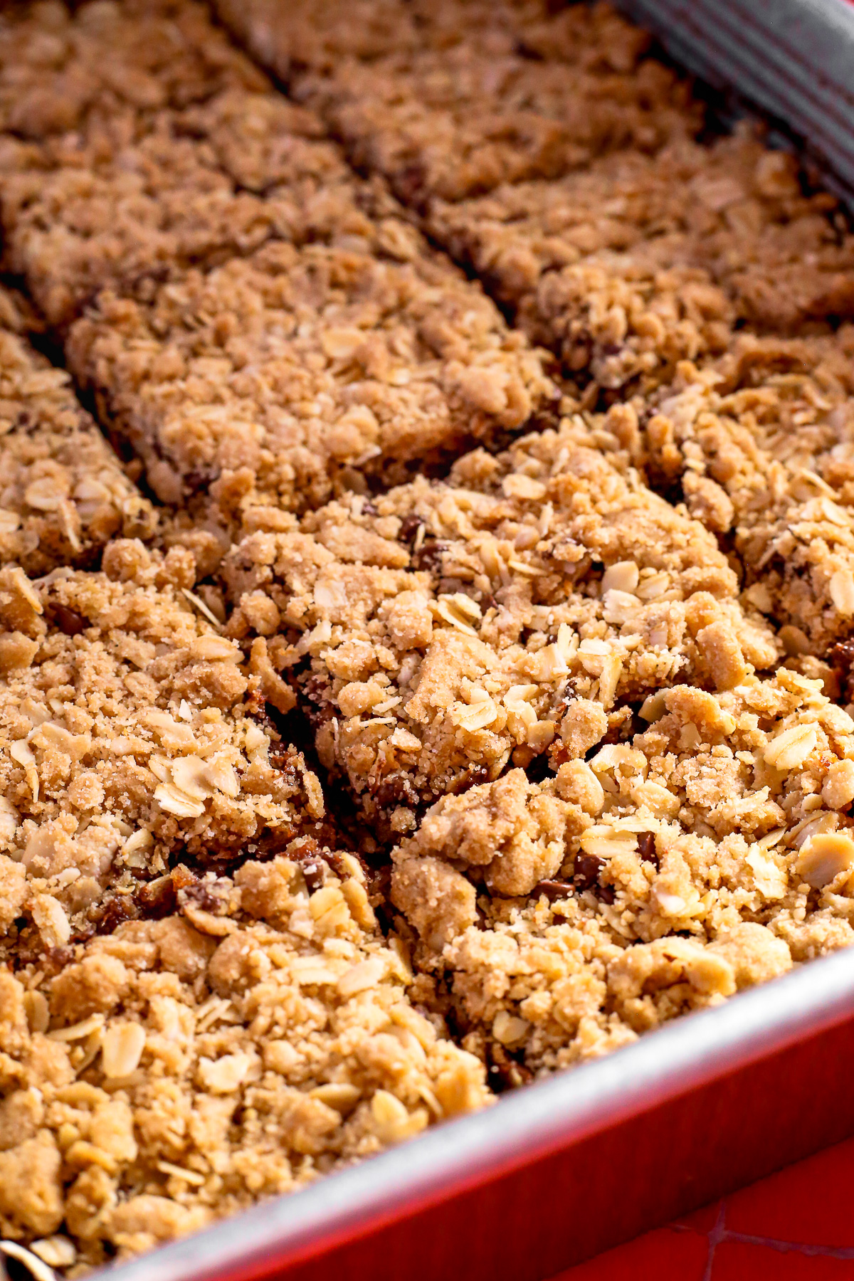 A baking pan full of freshly baked old fashioned date bars cut into squares, with a crumbly oat topping and melted chocolate chips poking through the top in some sections. 