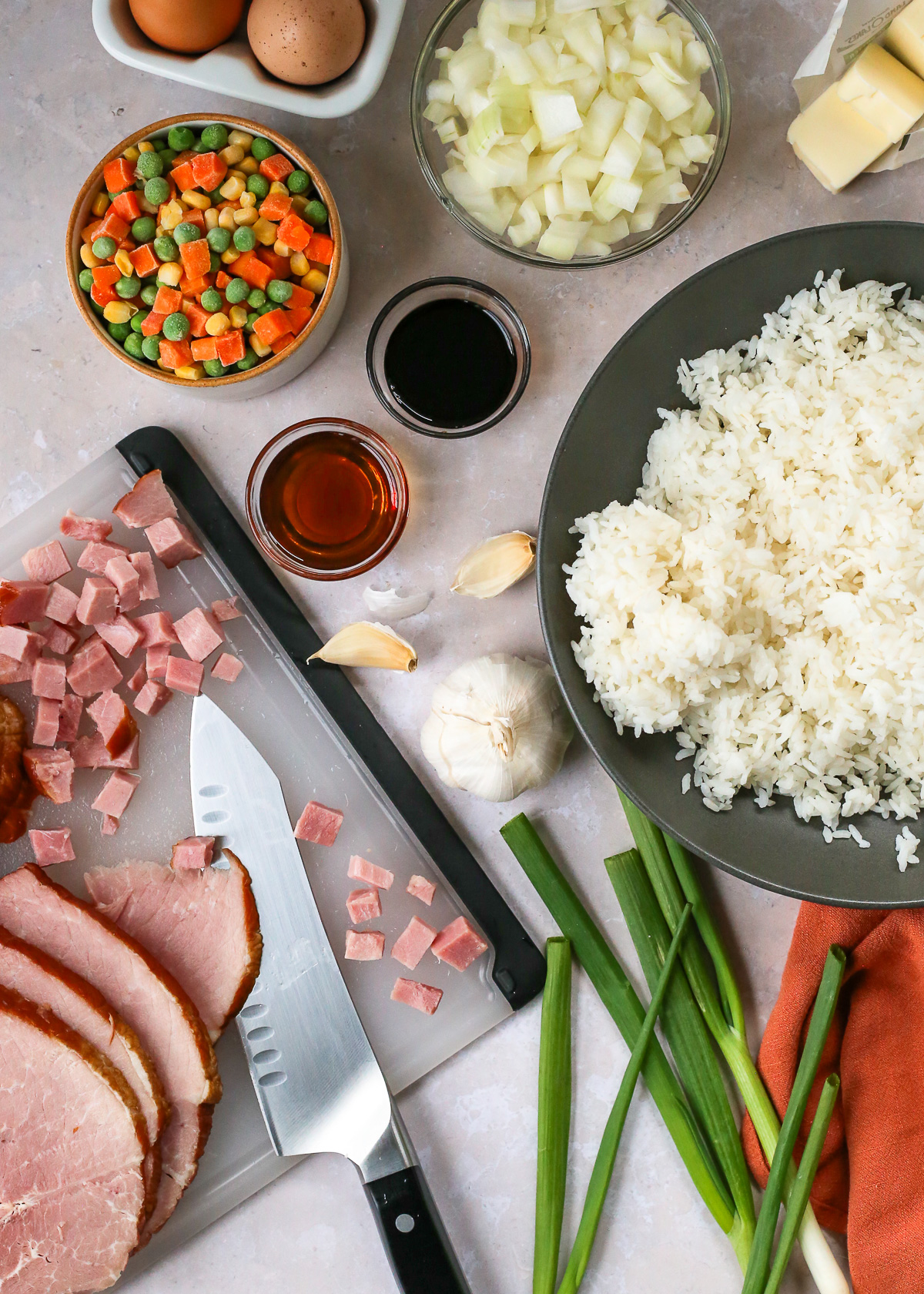All the ingredients displayed for a leftover ham fried rice recipe, including sliced and chopped ham, sesame oil, soy sauce, cooked white rice, green onions, diced onions, frozen mixed vegetables included corn, peas, and carrots, eggs, butter, and garlic.