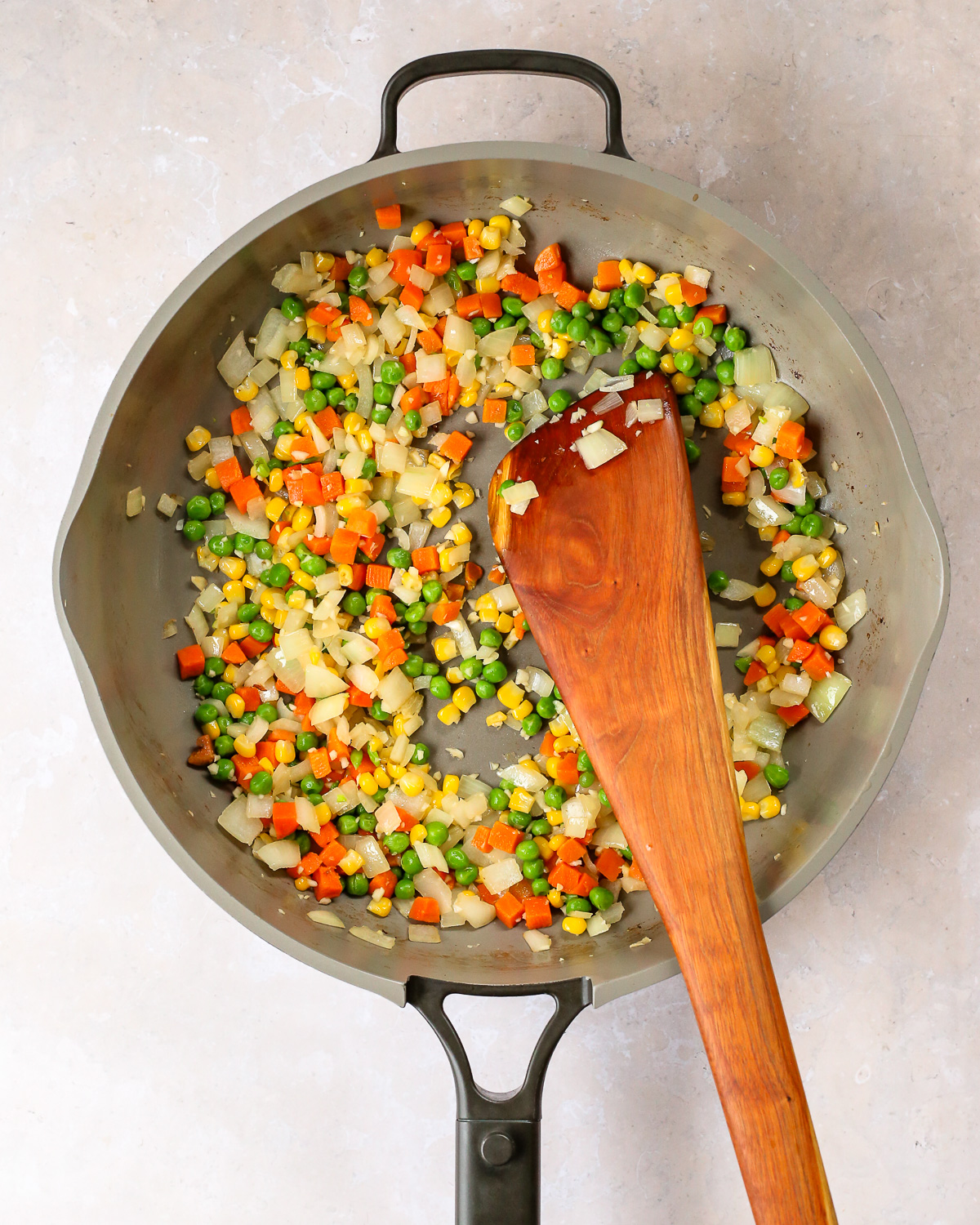 A wooden spatula stirs a vegetable mixture of onions, frozen peas, carrots, and corn in a large skillet.