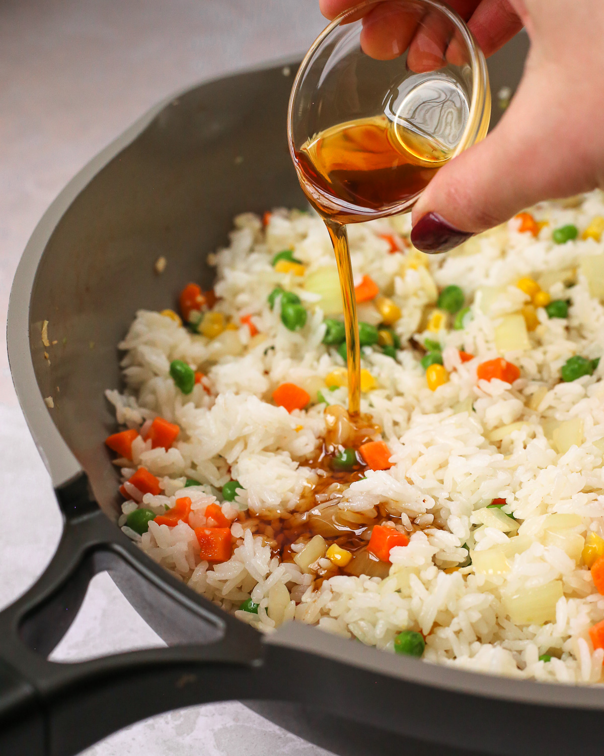 A woman's hand pours sesame oil from a small clear glass ramekin over a skillet full of fried rice.