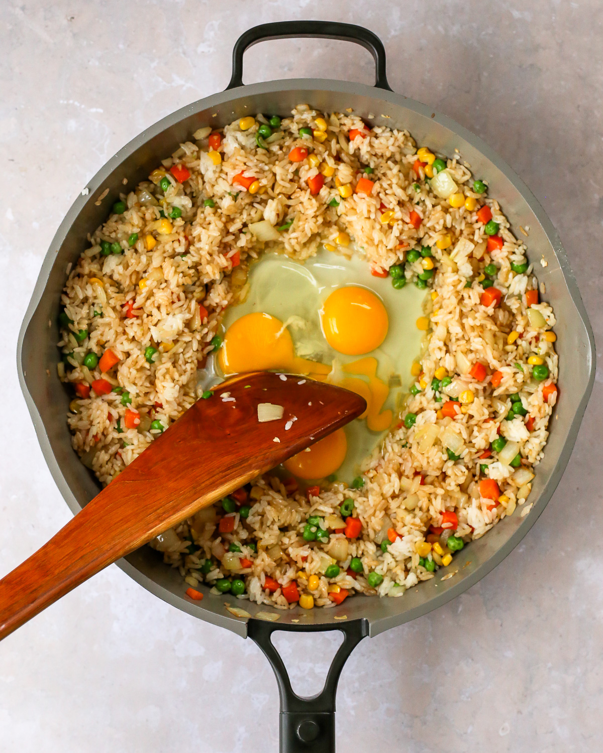 Three eggs are cracked into an open well in the center of a large skillet used to cook a ham fried rice recipe, with a wooden spatula resting on the side to cook the scrambled eggs.