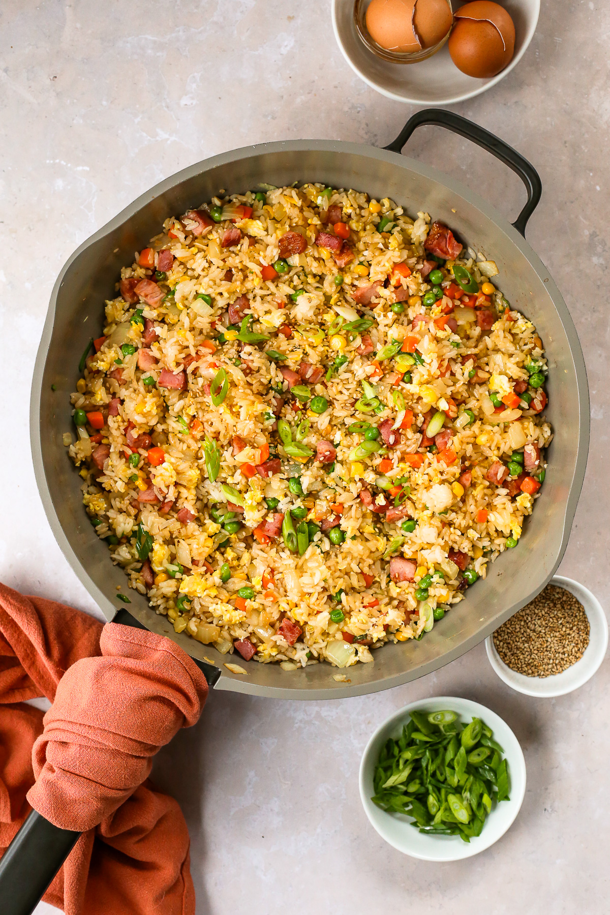 Overhead view of a large skillet full of ham fried rice, with small ramekins for sliced green onions and toasted sesame seeds on a kitchen countertop.