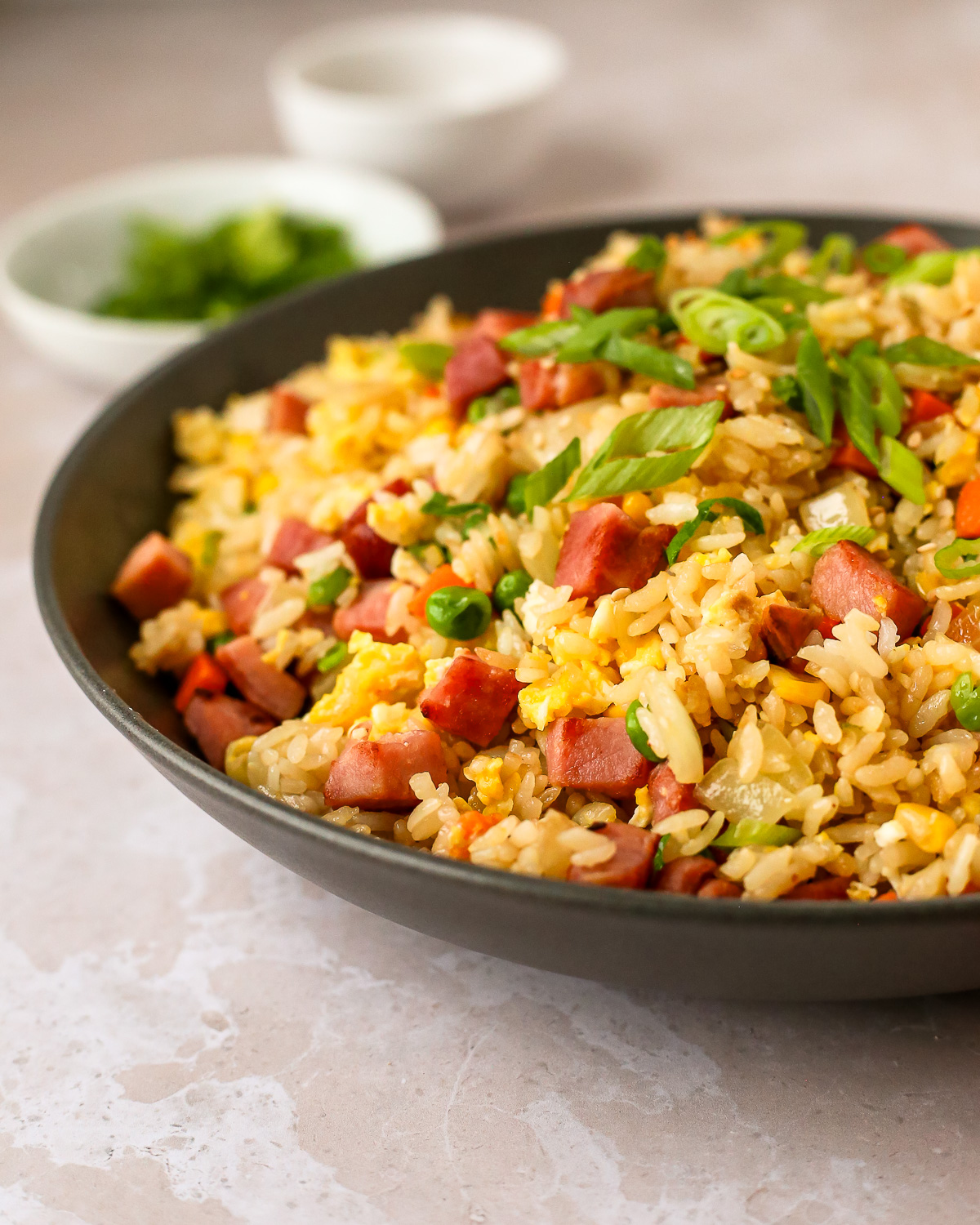 A large serving bowl full of ham fried rice displayed on a kitchen countertop with sliced green onions for garnish in the background.