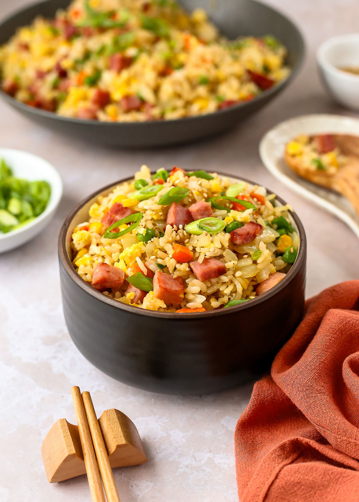A ceramic bowl filled with ham fried rice, served on a kitchen countertop with a pair of wooden chopsticks and a faded orange linen napkin.