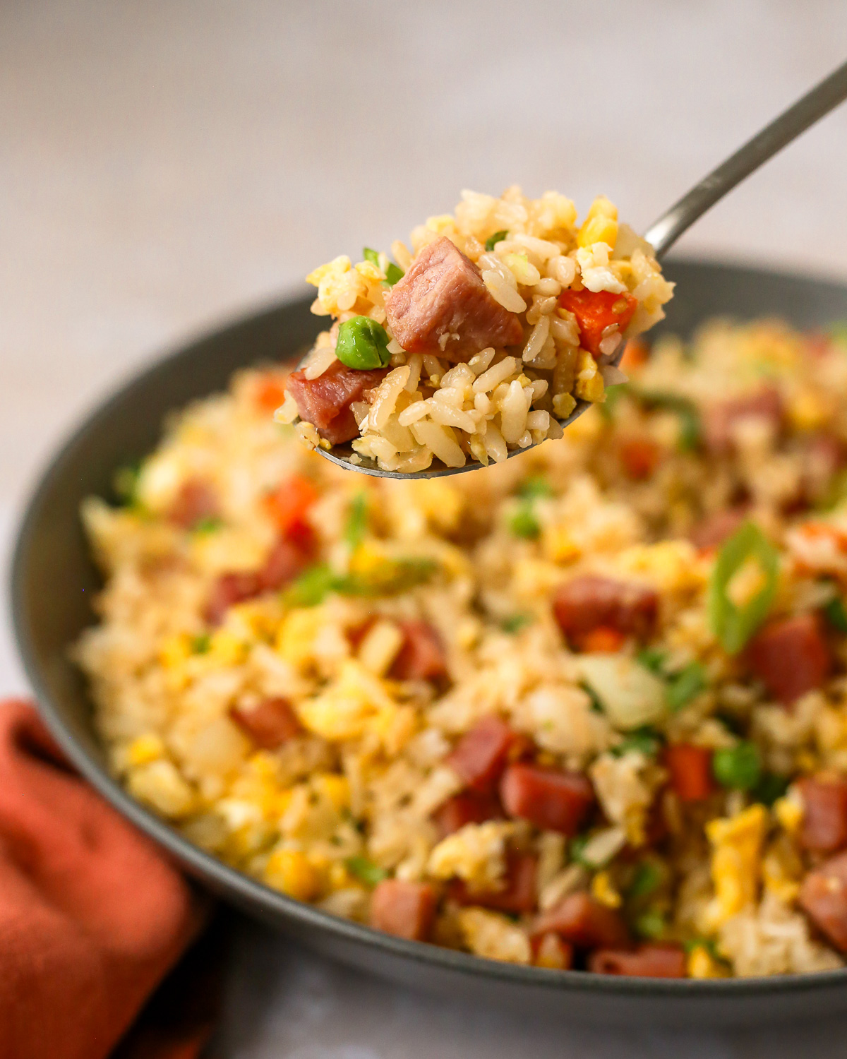 A silver serving spoon lifts a scoop of ham fried rice from a serving bowl, showing the grains of rice mixed with seared ham, vegetables, and small pieces of scrambled eggs.