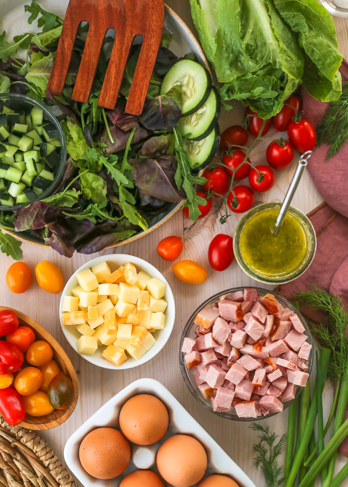 An assortment of ingredients for a chefs salad recipe, including a carton of brown eggs, cherry tomatoes, chopped ham and cheese, fresh dill and green onions, romaine lettuce, and diced cucumbers displayed on a kitchen table.