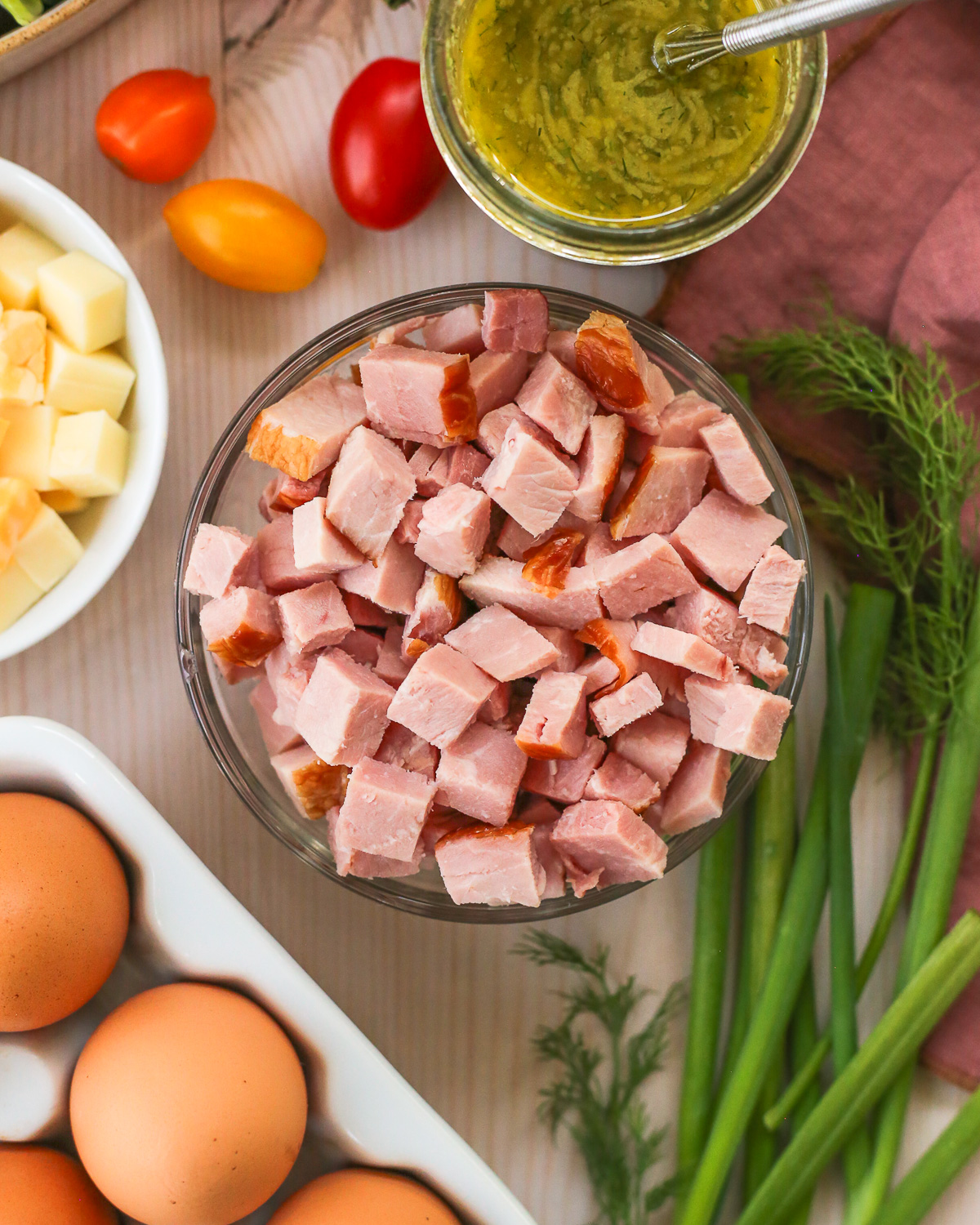 Overhead view of a small glass prep bowl filled with chopped ham, surrounded by the ingredients to make a leftover ham recipe.